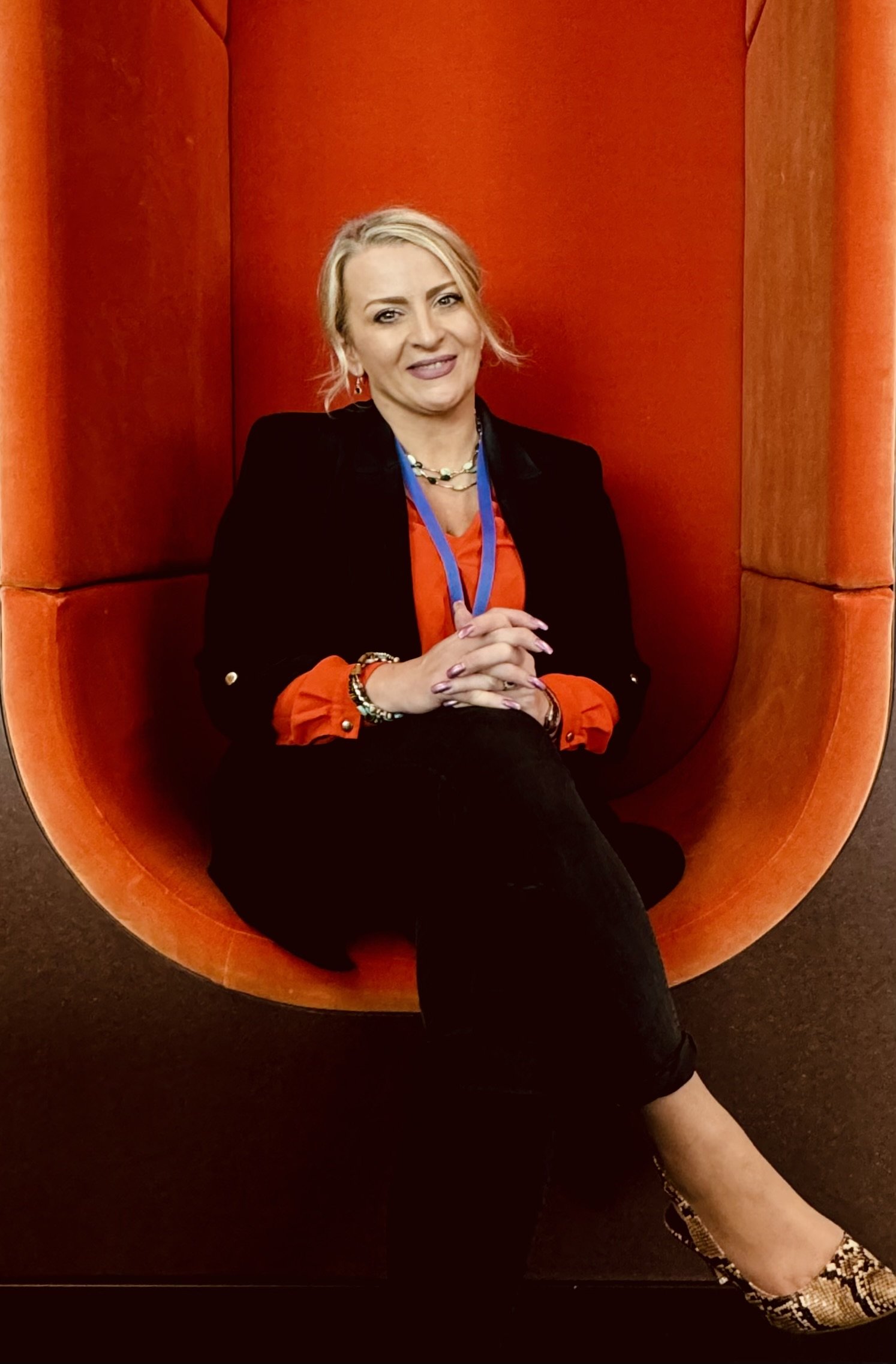 A woman with blonde hair sits in an orange, egg-shaped chair. She is wearing a black blazer, an orange blouse, black pants, patterned shoes, and jewelry, including a necklace and bracelets. She has a blue lanyard around her neck and is smiling at the camera.
