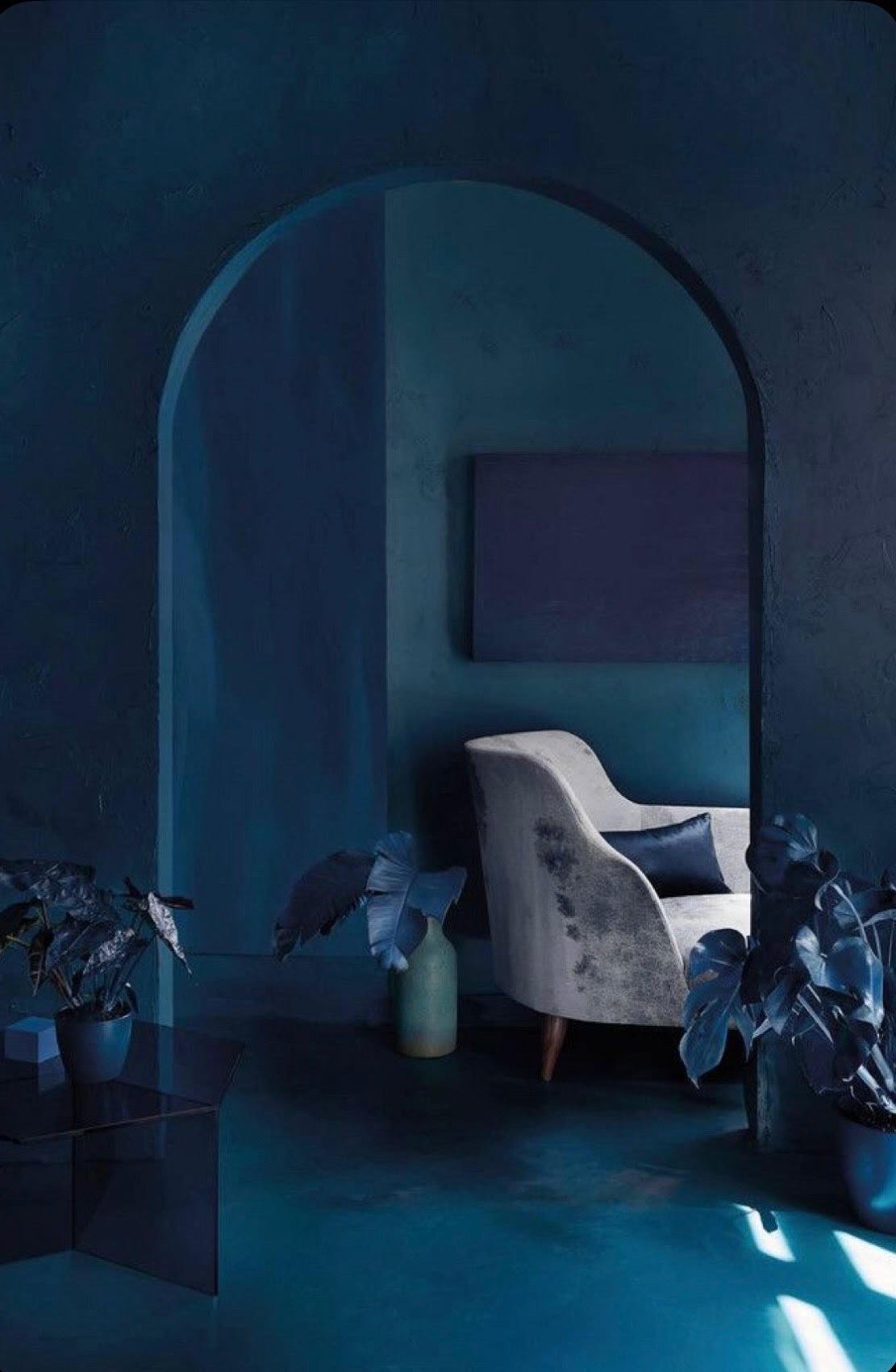 A cozy interior with dark blue walls and a light gray armchair, accented by plants in blue pots, a black geometric coffee table, and minimalist decor.