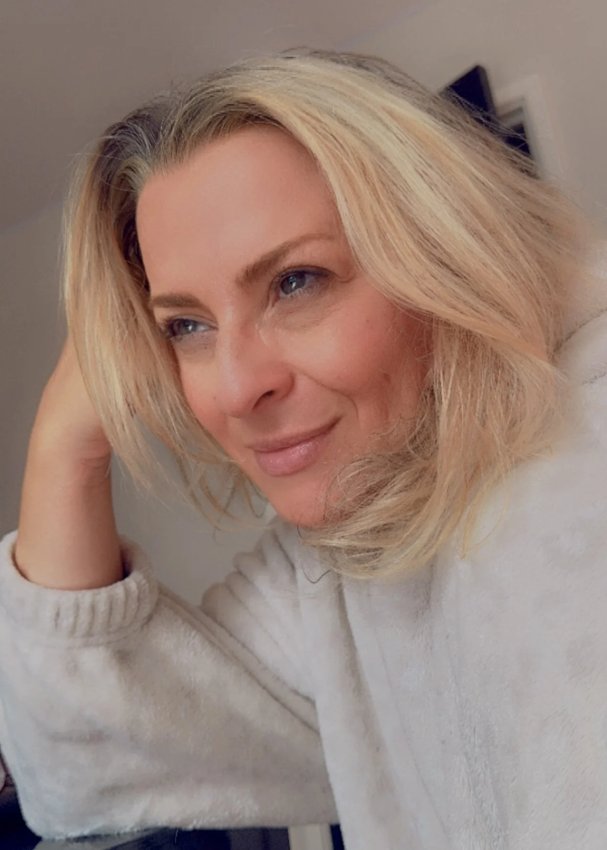 Close-up of a woman with blonde hair, blue eyes, smiling softly, resting her head on her hand, wearing a white sweater.