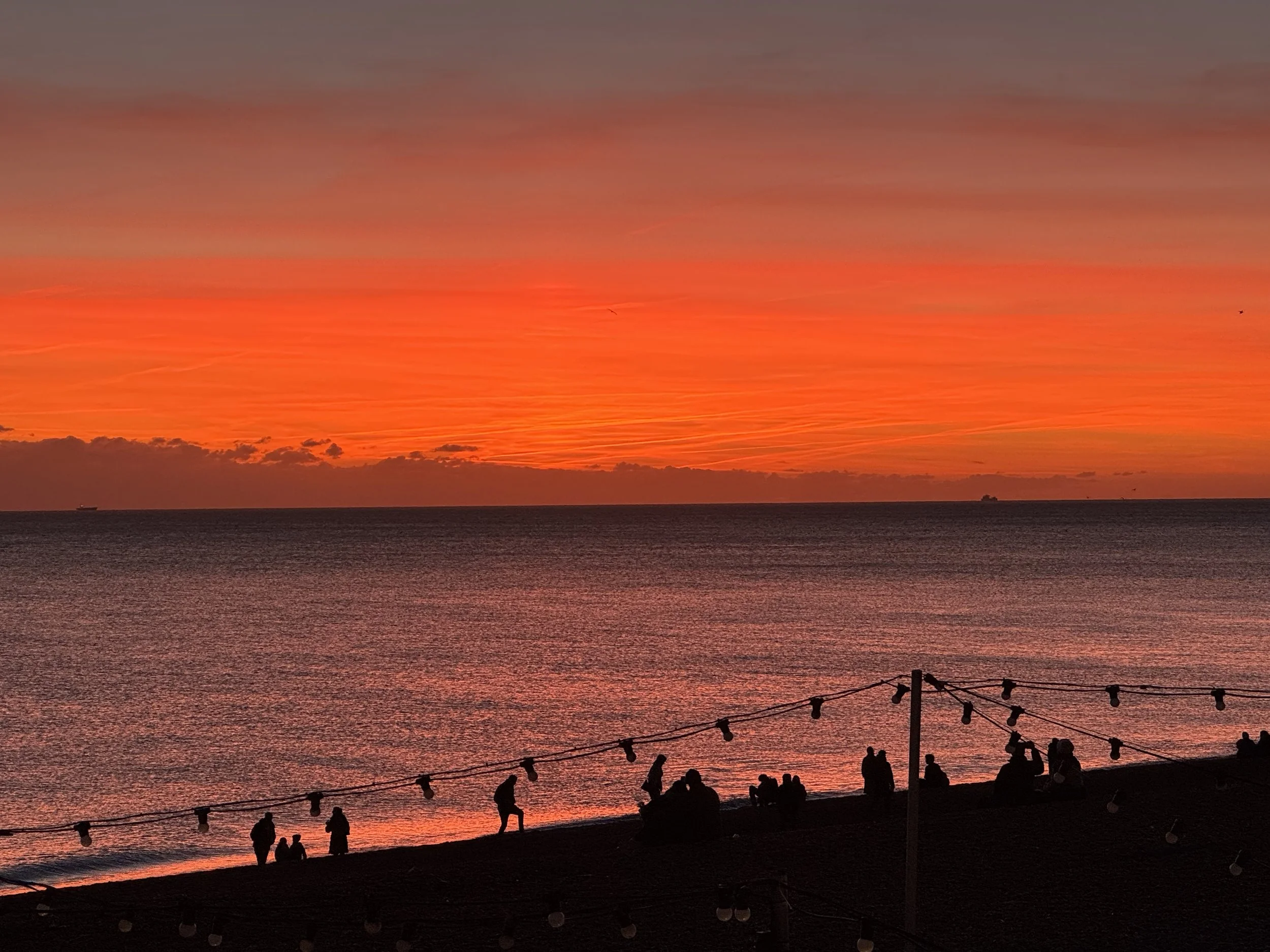 People walking and sitting on a beach at sunset with vibrant orange and pink sky, string lights hanging above.