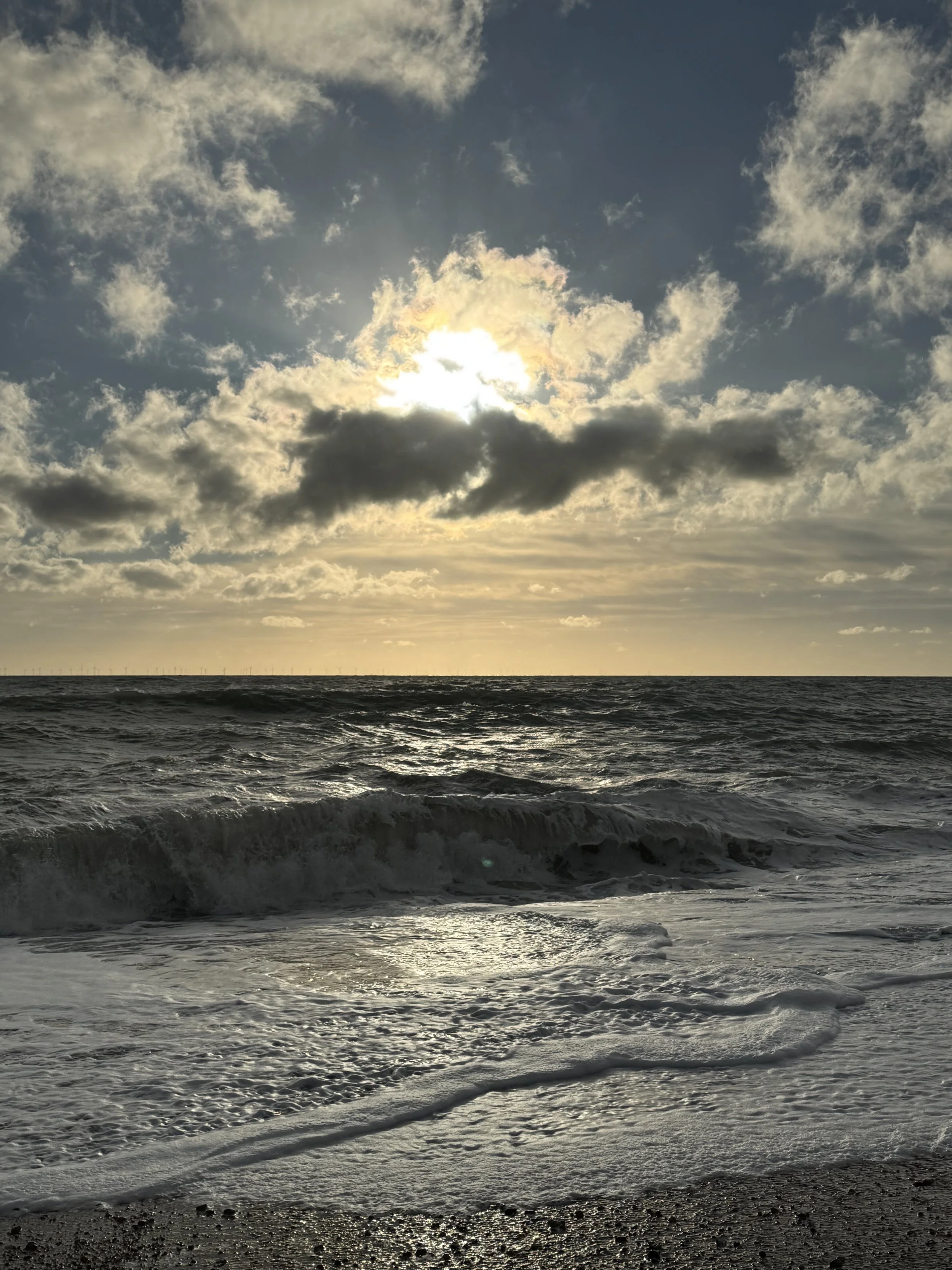 Sun peeking through clouds over the ocean with waves crashing on the shore at sunset.