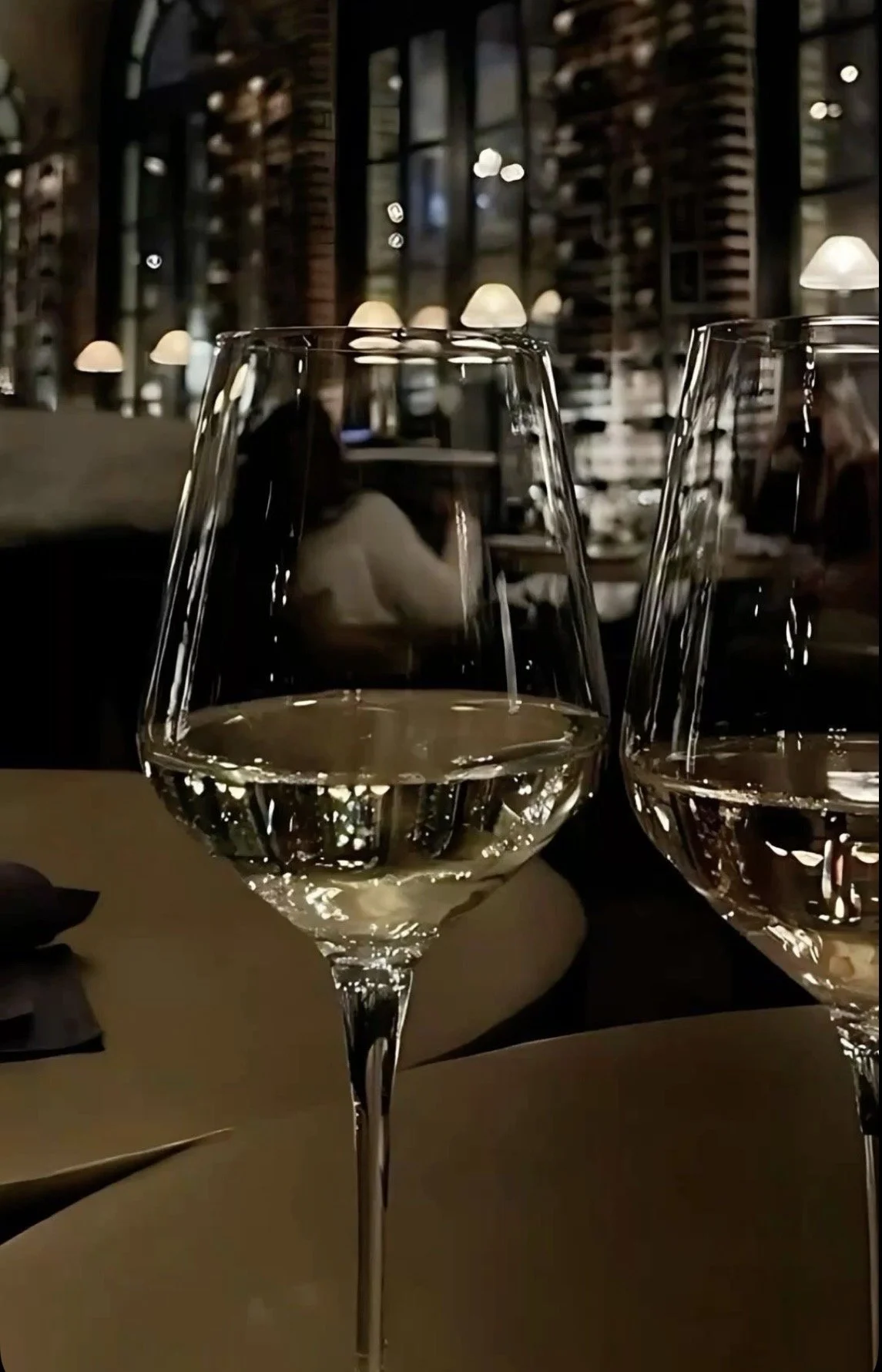Two glasses of white wine on a table at a restaurant with warm lighting and shelves filled with books in the background.