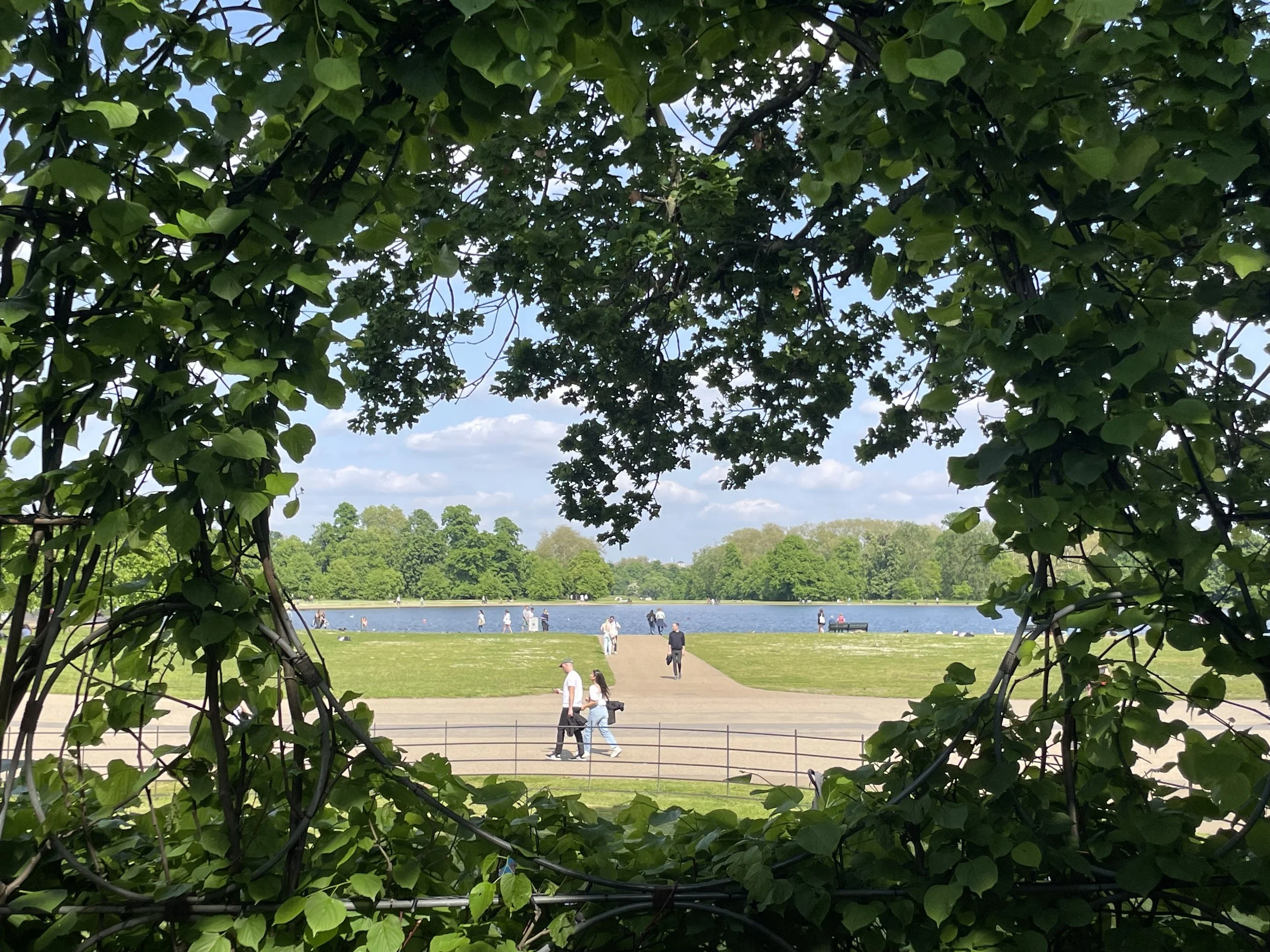 View of a park with a lake in the distance, framed by green leaves and branches in the foreground, with people walking along the path and by the water on a sunny day.
