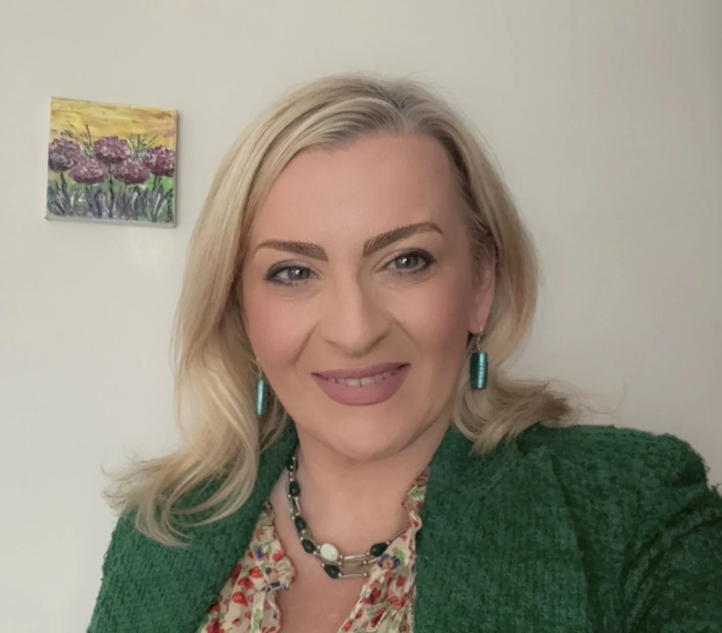 A woman with blonde hair wearing a green jacket, earrings, necklace, and floral top, smiling in front of a plain wall with a colorful painting of flowers.
