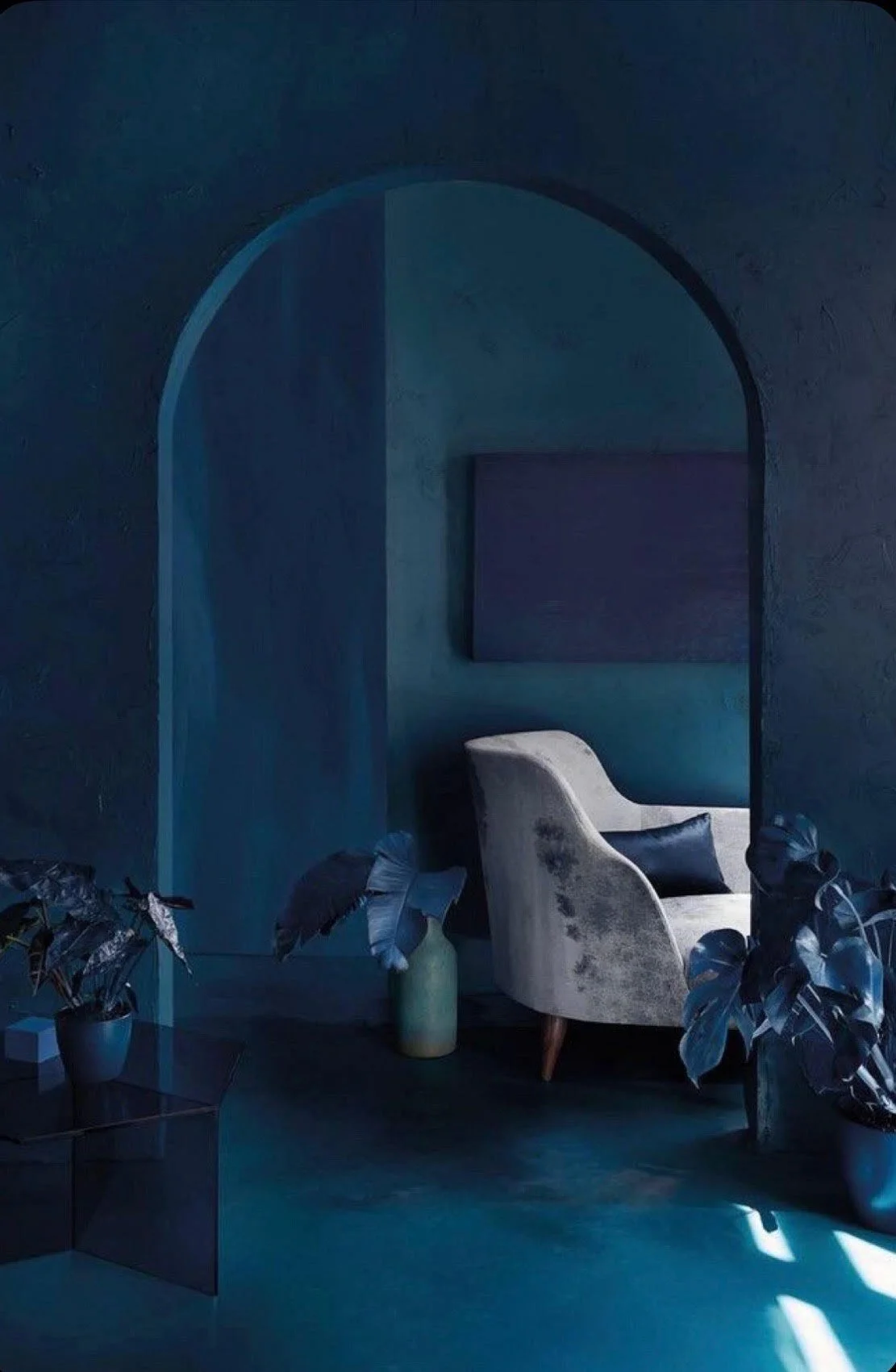 A cozy corner in a dark blue room with a plush armchair, some plants, and abstract artwork.