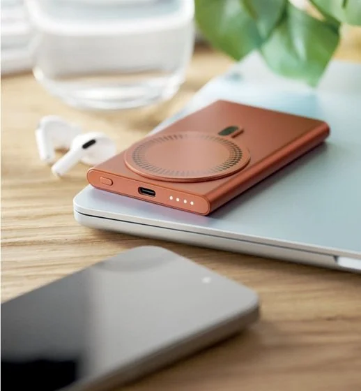 A flat lay of a smartphone with a wireless charging pad on top, a closed laptop, a glass of water, and a pair of wireless earbuds on a wooden desk with a plant in the background.