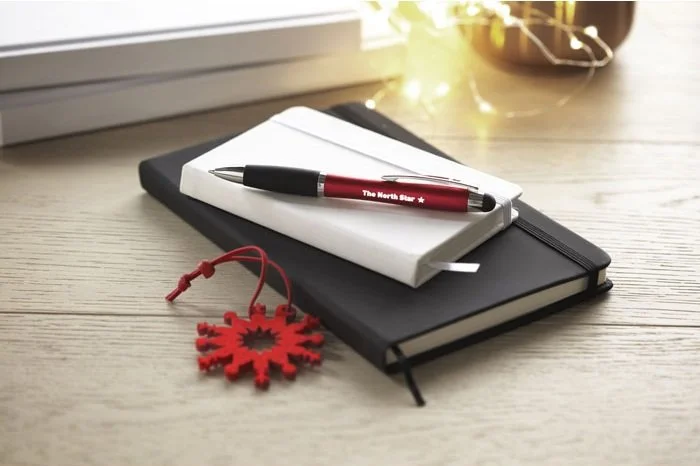 Desk with black notebook, white notepad, red pen, and red snowflake-shaped holiday ornament on light wood surface.