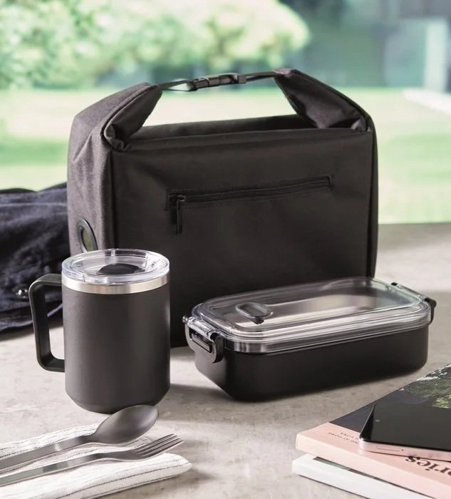 A black insulated lunch bag, a black travel mug with a clear lid, a black food container with a clear lid, a fork, a spoon, a napkin, some books, and a smartphone on a table against a window showing green outdoors.