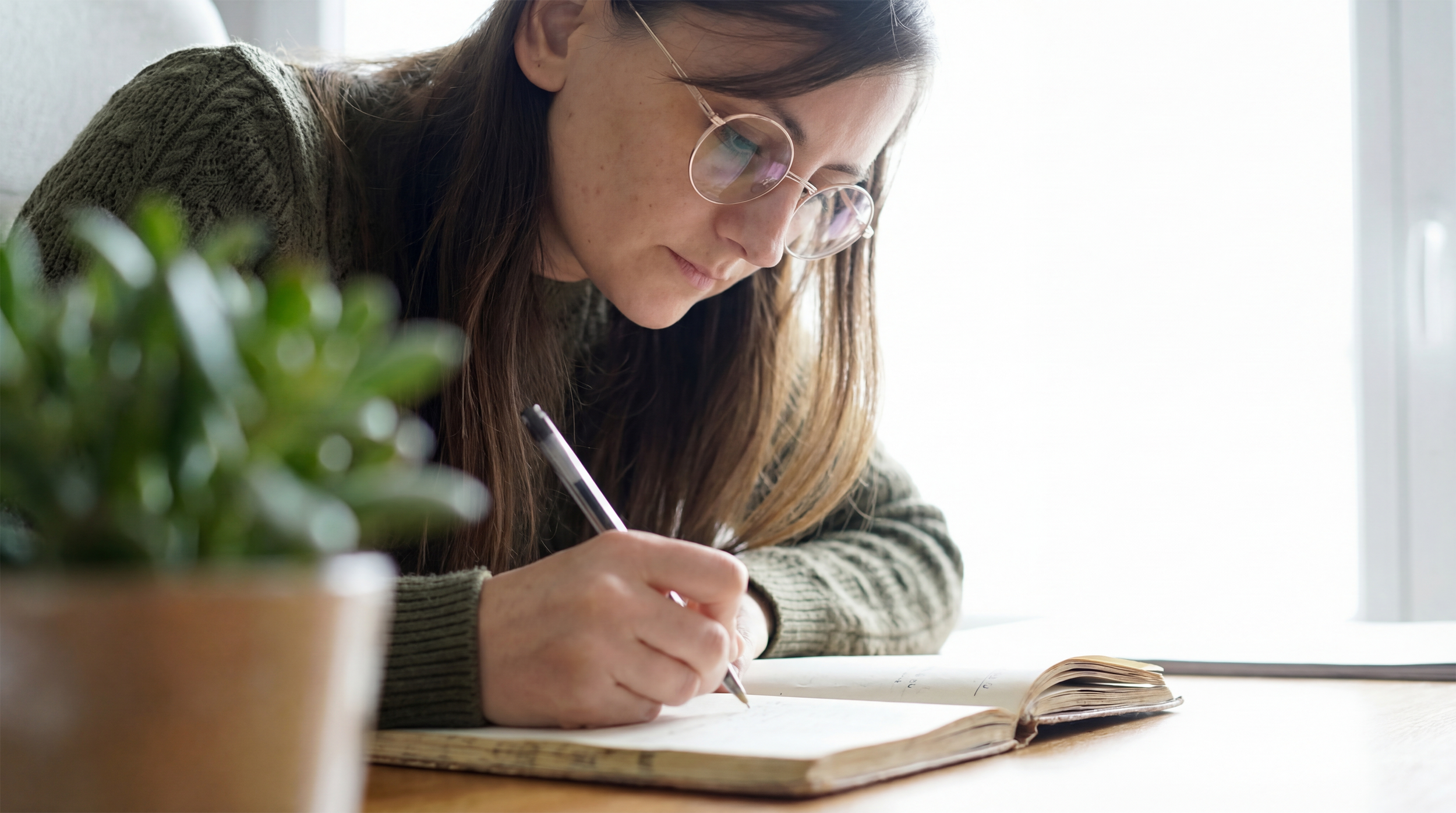 Une femme avec des lunettes et un pull en laine écrivant dans un carnet posé sur une table, à proximité d'une plante verte en pot, près d'une fenêtre lumineuse.