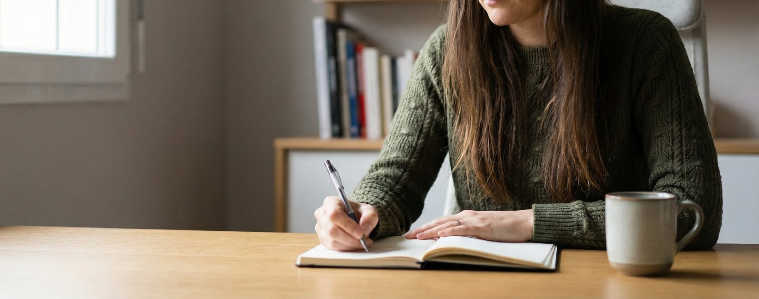 Lauriane Celton Pénisson assise à une table, écrivant dans un carnet avec une tasse à café à côté, intérieur d'un bureau, bibliothèque en arrière-plan.