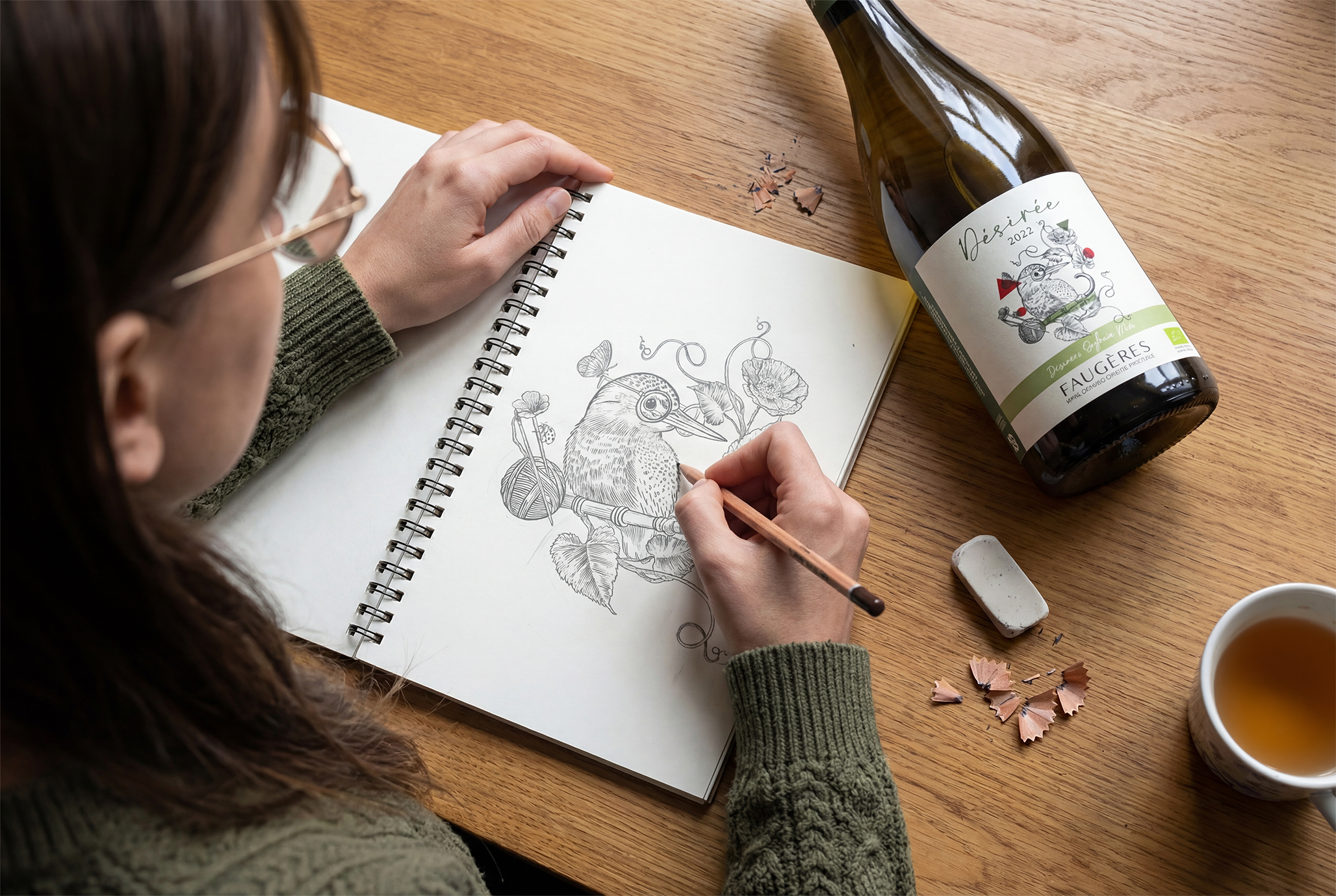 Une femme dessine un oiseau sur un carnet à croquis, avec un stylo, sur une table en bois, entourée d'une bouteille de vin, d'un mug de thé, de débris de crayons de couleur, et d'une gomme.