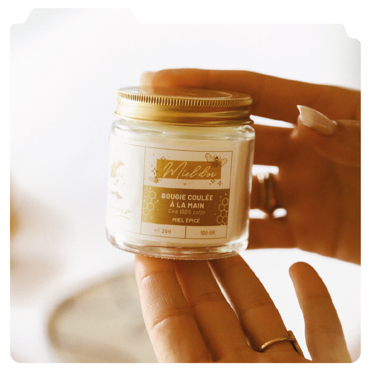 Une main tient un pot de bougie faite à la main à base de cire 100% de colza, au parfum de miel épicé, avec un couvercle doré.