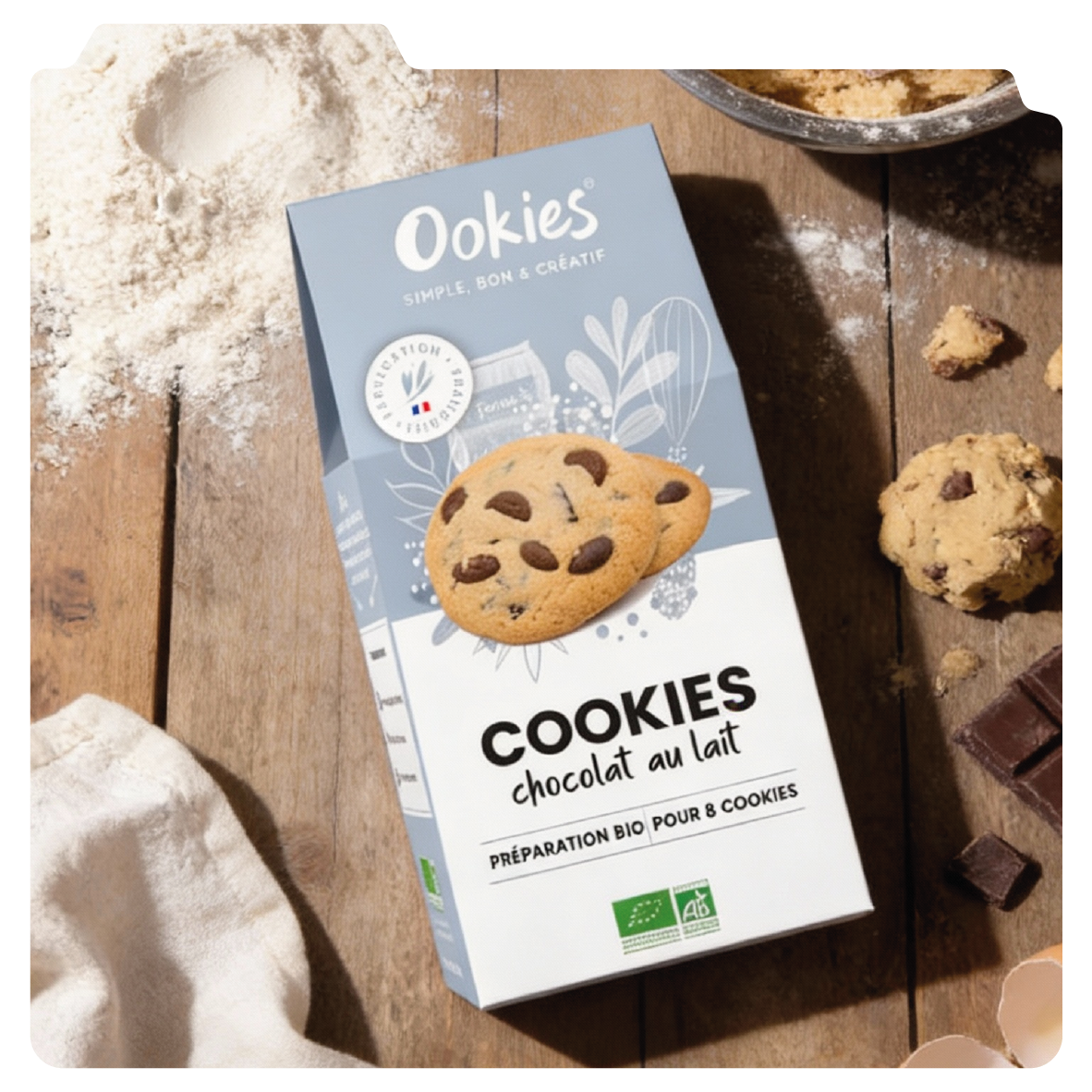 Design graphique d'un packaging de préparation de cookies au chocolat au lait. La boîte est bleue et blanche avec une image de cookies aux pépites de chocolat. Il y a des morceaux de cookies autour et un peu de farine sur une table en bois.