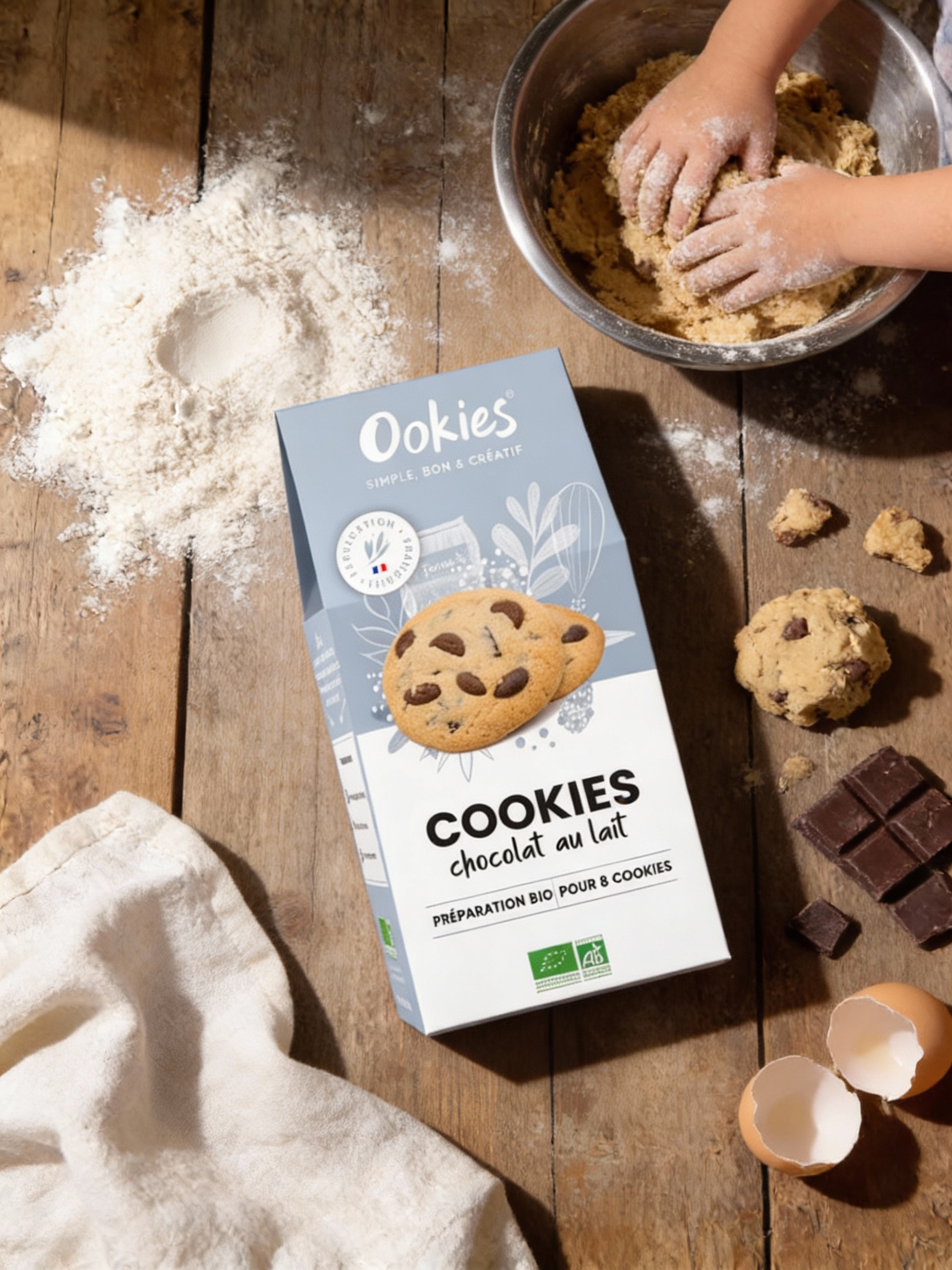 Design graphique d'un packaging de préparation pour cookies au chocolat au lait, à côté de morceaux de chocolat, de la farine, des œufs cassés, et quelques cookies crues sur une table en bois, avec des mains d'enfant en train de pétrir la pâte.