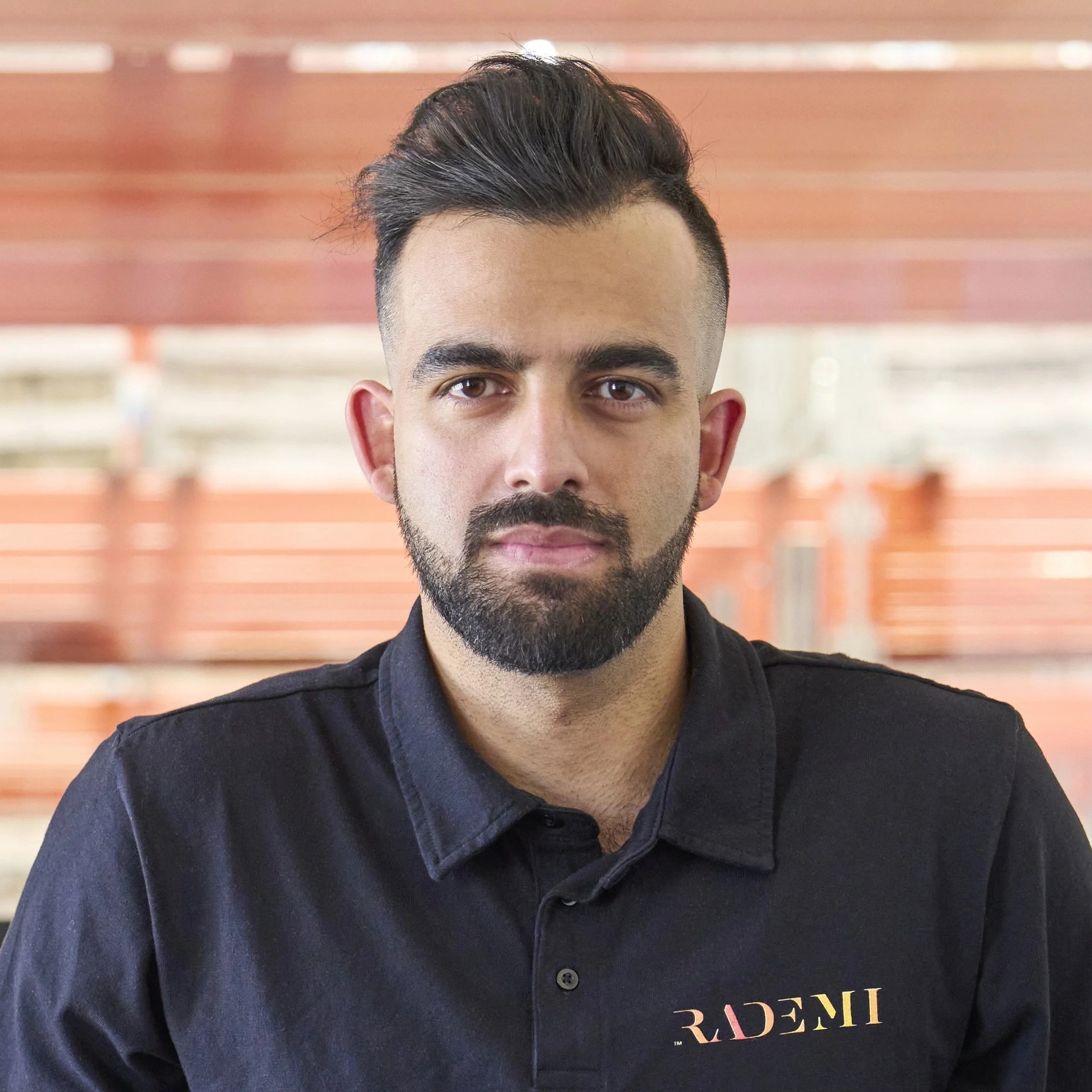 A man with dark hair, a beard, and a slight mustache, wearing a black shirt with the logo 'RADENI' on it, standing indoors with blurred wooden shelves in the background.