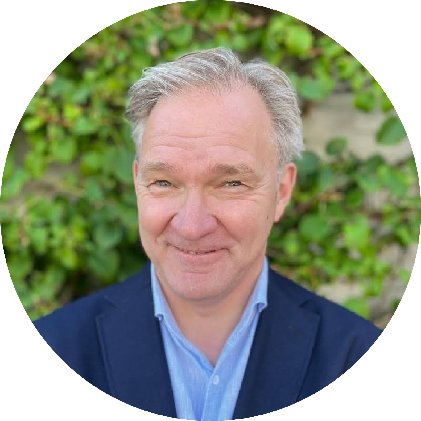 Headshot of a middle-aged man with gray hair, smiling, wearing a blue blazer and light blue shirt, outdoors with green foliage in the background.