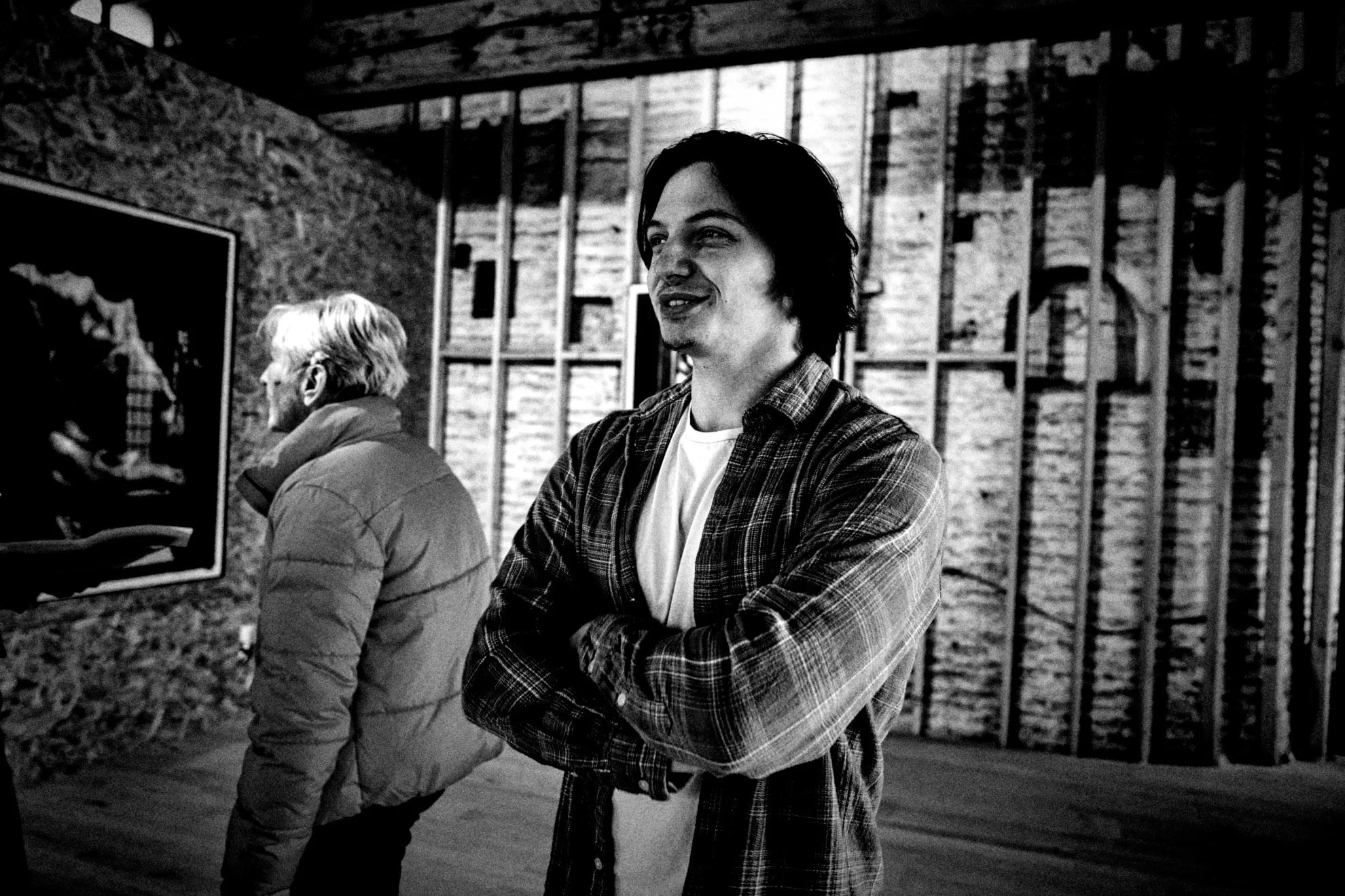 Two men in an art gallery, with one in the foreground smiling and crossing his arms, wearing a plaid shirt, and the other in the background looking at artwork, wearing a puffer jacket. The walls are wooden with exposed beams.