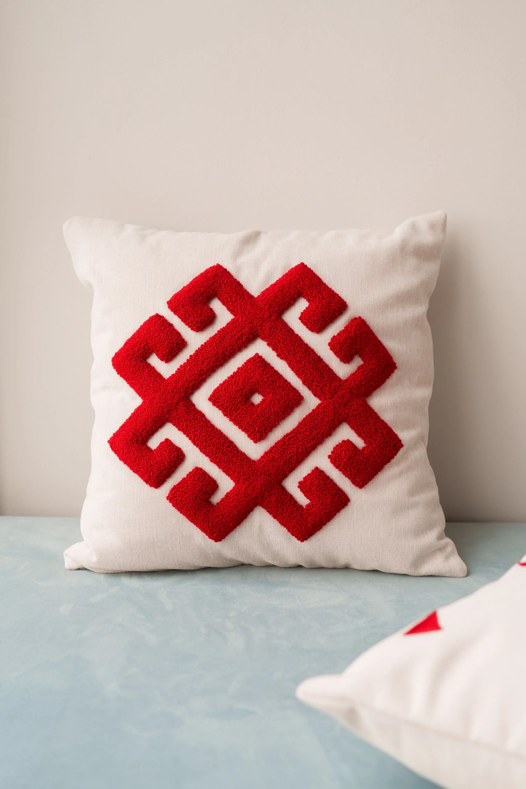A white decorative pillow with a red geometric pattern, placed on a bed with a light blue cover.