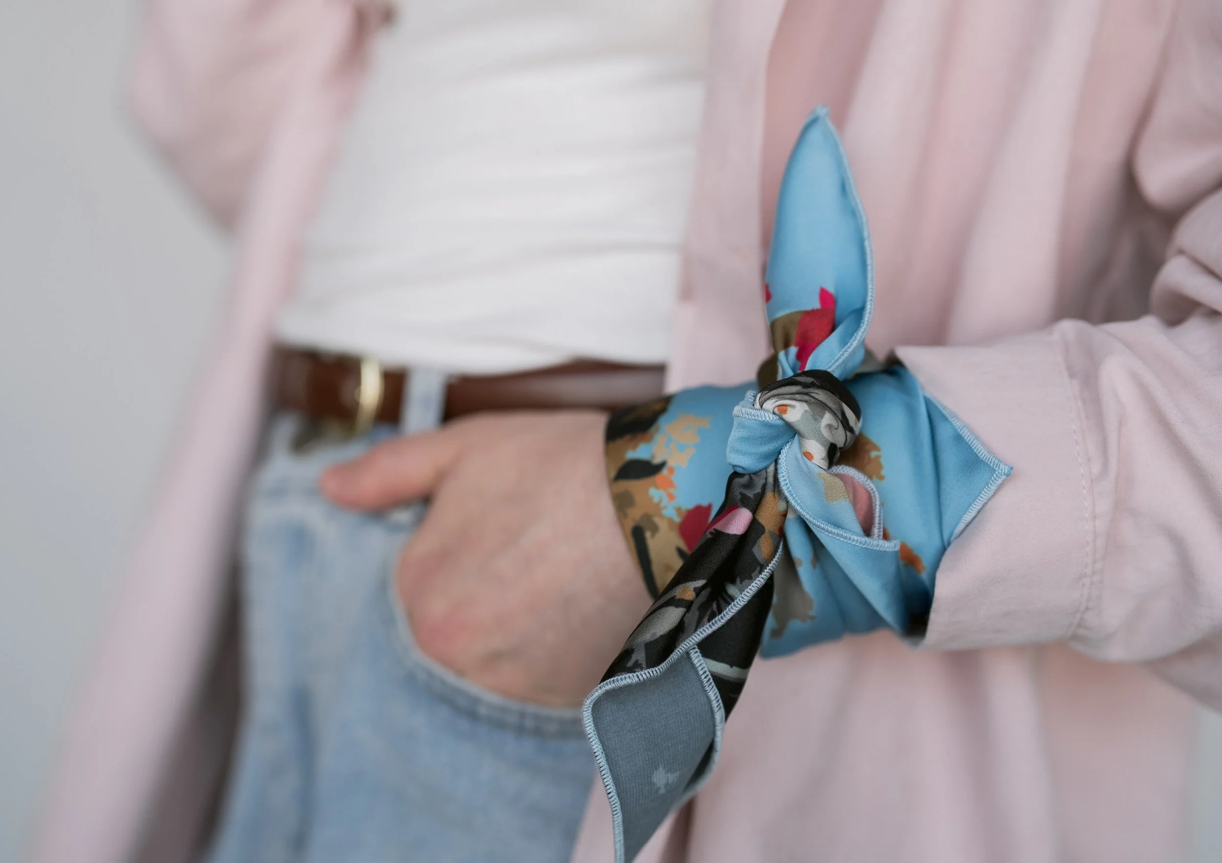Close-up of a person's wrist with a colorful silk scarf tied in a knot, with their hand resting on their hip, wearing light blue jeans and a light pink blazer.