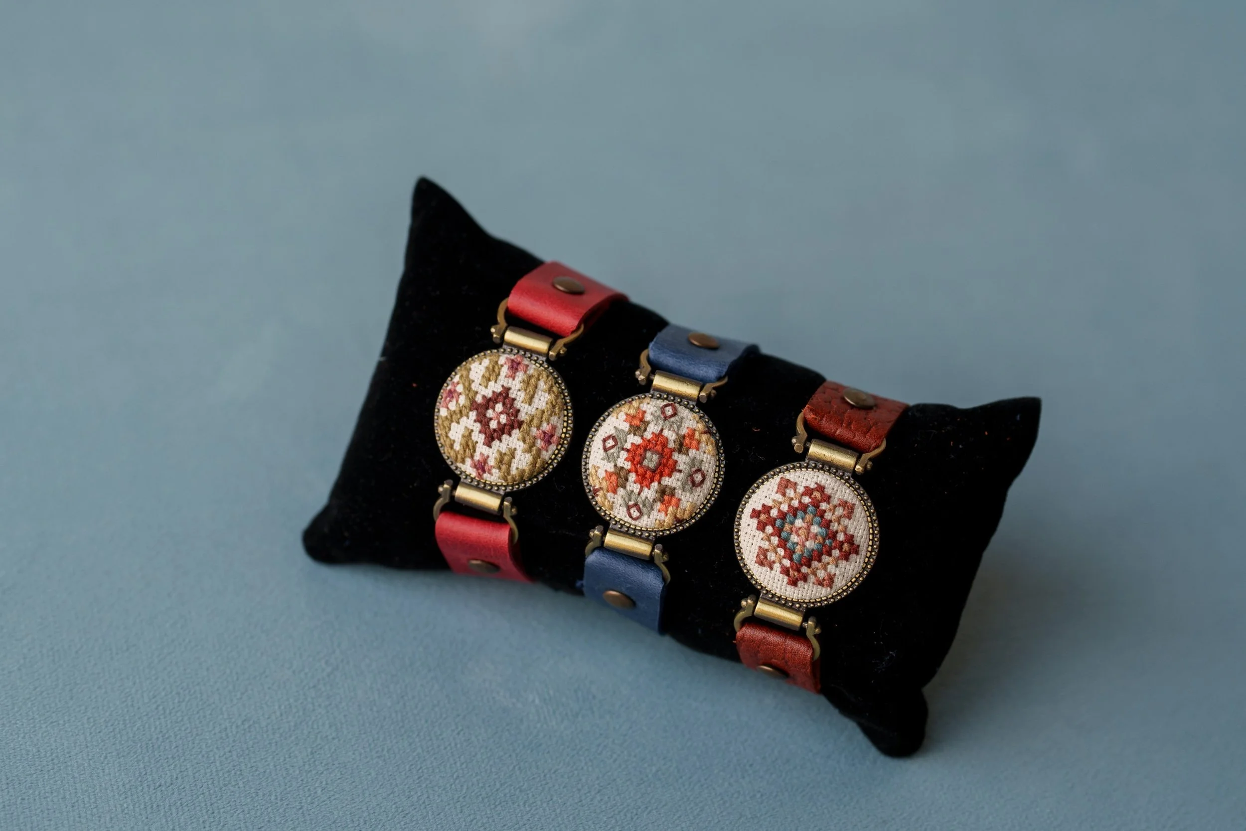 Three embroidered circular patches with floral patterns, each attached to leather straps in red, blue, and red colors, displayed on a black velvet pillow.