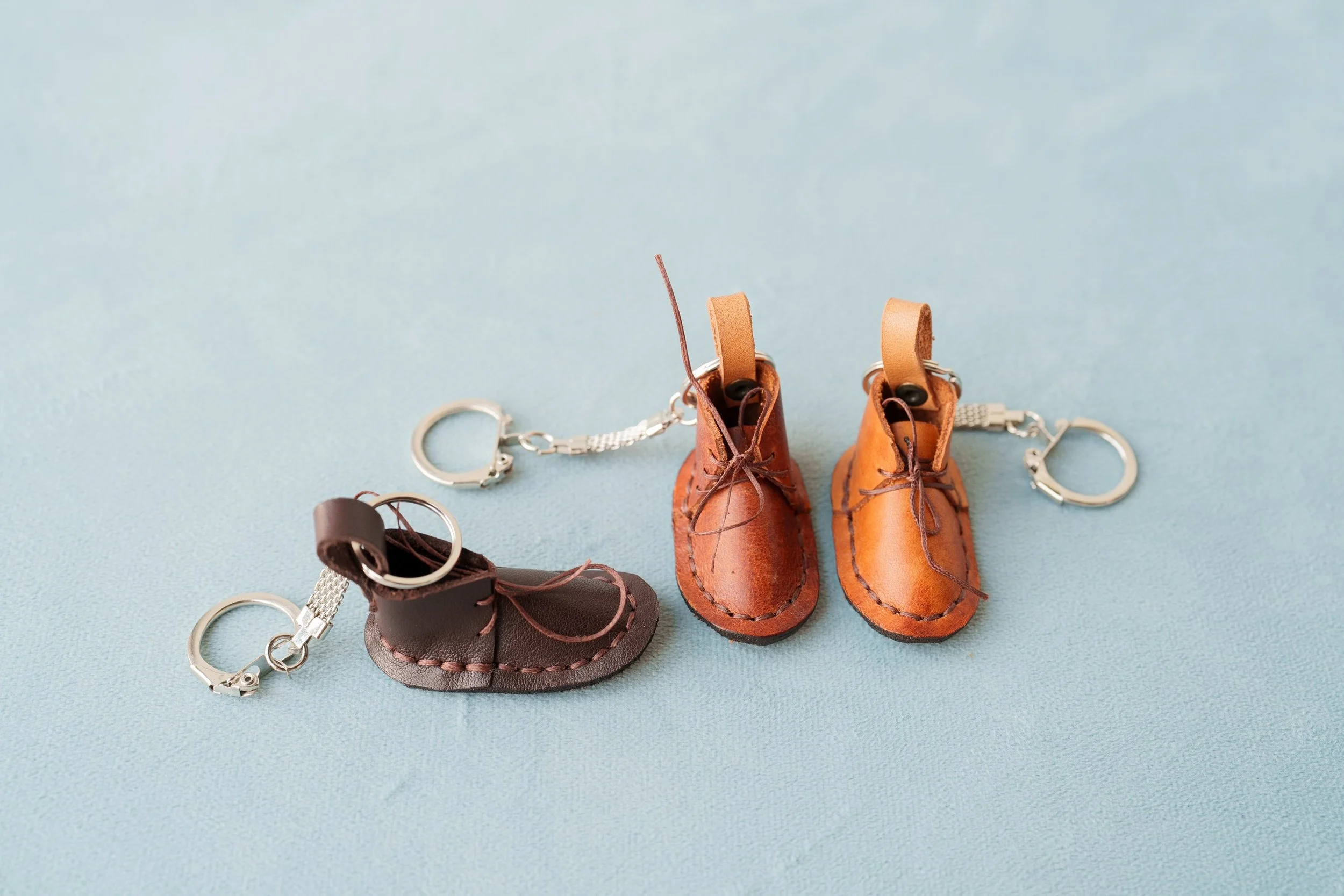 Miniature leather shoes with keychains attached, one dark brown and two light brown, on a light blue surface.