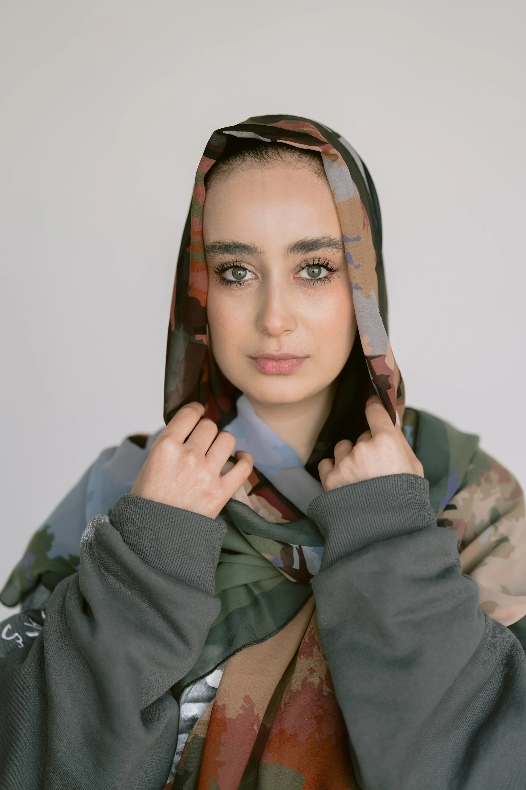 A woman with fair skin and green eyes, wearing a camouflage-patterned hooded garment and a gray hoodie, looks directly at the camera with a neutral expression.