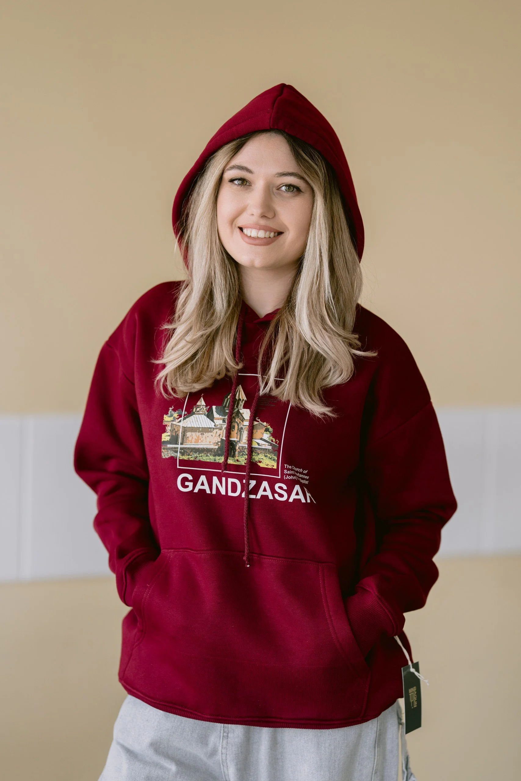 A young woman smiling, wearing a red hooded sweatshirt with the word 'GANDZASAR' and a graphic of a church, standing indoors against a beige and white background.