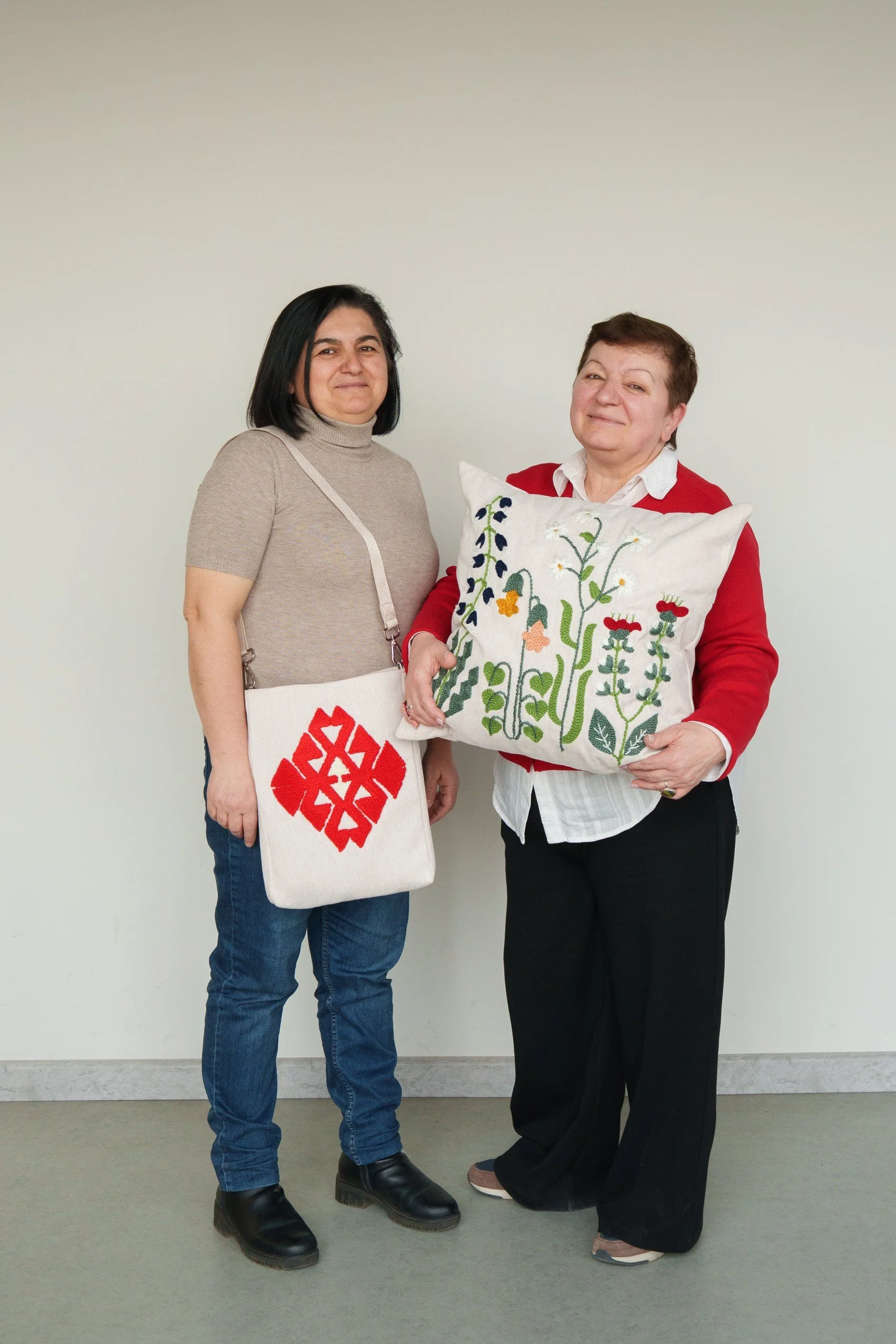 Two women standing against a plain wall, one holding a decorative pillow with embroidered plants and flowers, and the other wearing a shoulder bag with a red geometric design.
