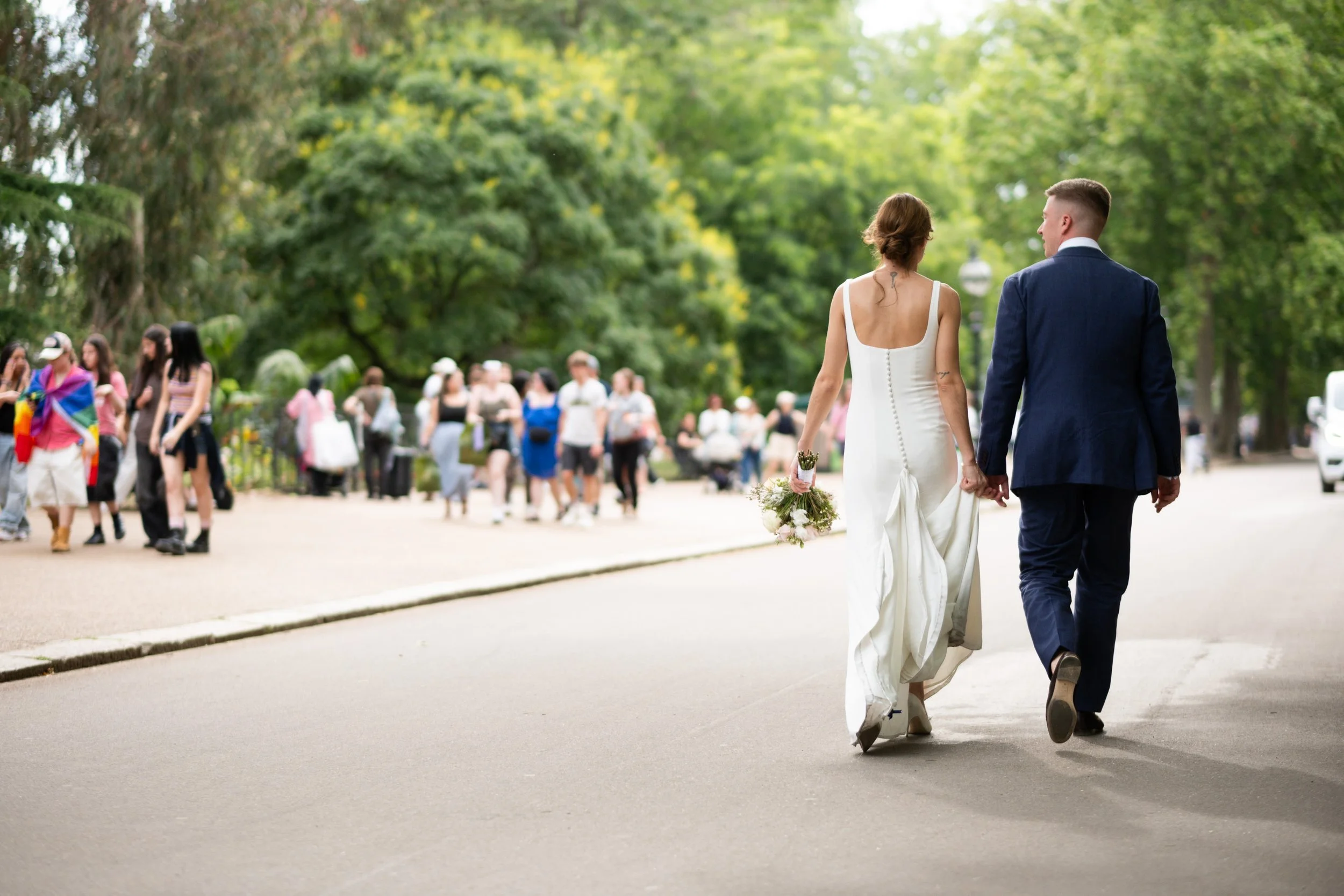 A bride and groom walking hand in hand on a street in a park, with people gathering in the background.