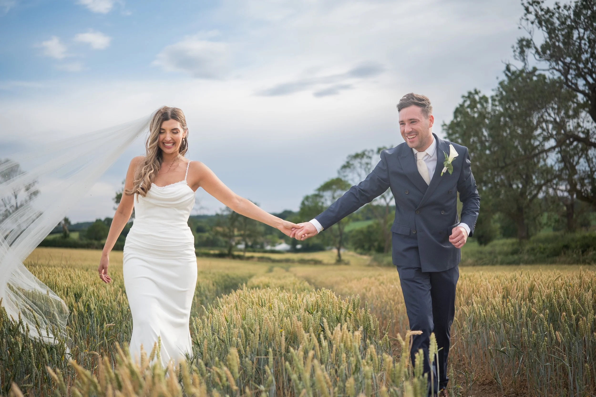 A bride and groom holding hands and smiling in a field of tall grass or wheat, with trees and a cloudy sky in the background.