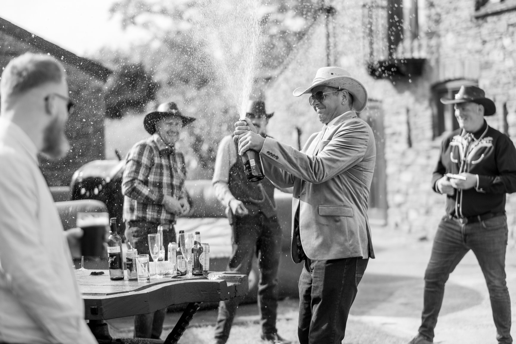 Man wearing cowboy hat and sunglasses opening champagne bottle at outdoor gathering with friends, some wearing cowboy hats and plaid shirts, near a rustic building.