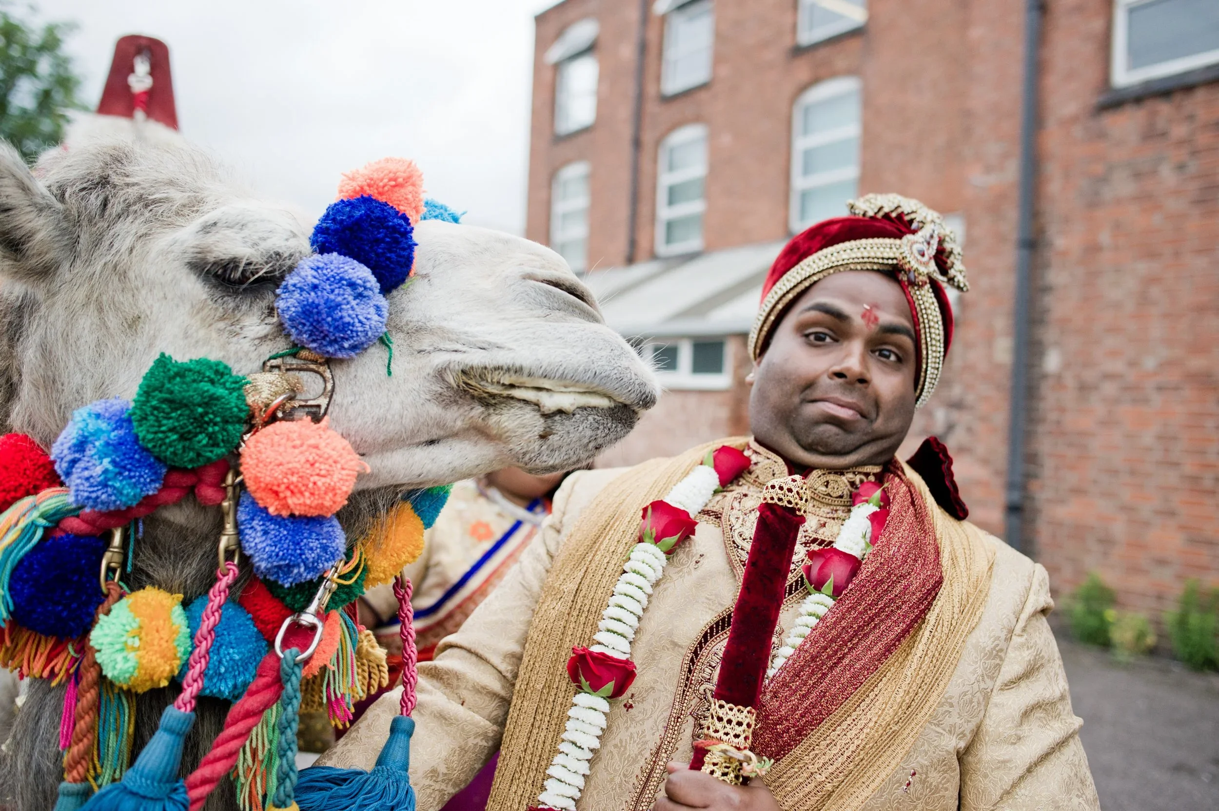A man dressed in traditional Indian wedding attire holding a ceremonial sword standing next to a decorated camel with colorful tassels and pom-poms.