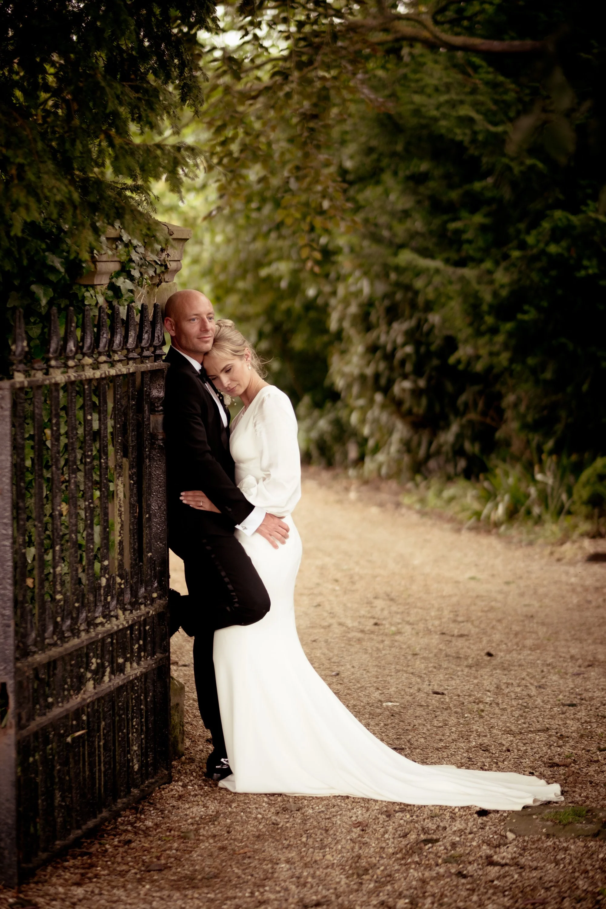 A bride and groom are standing close together, leaning against a black iron gate surrounded by greenery and trees, during daytime or early evening.