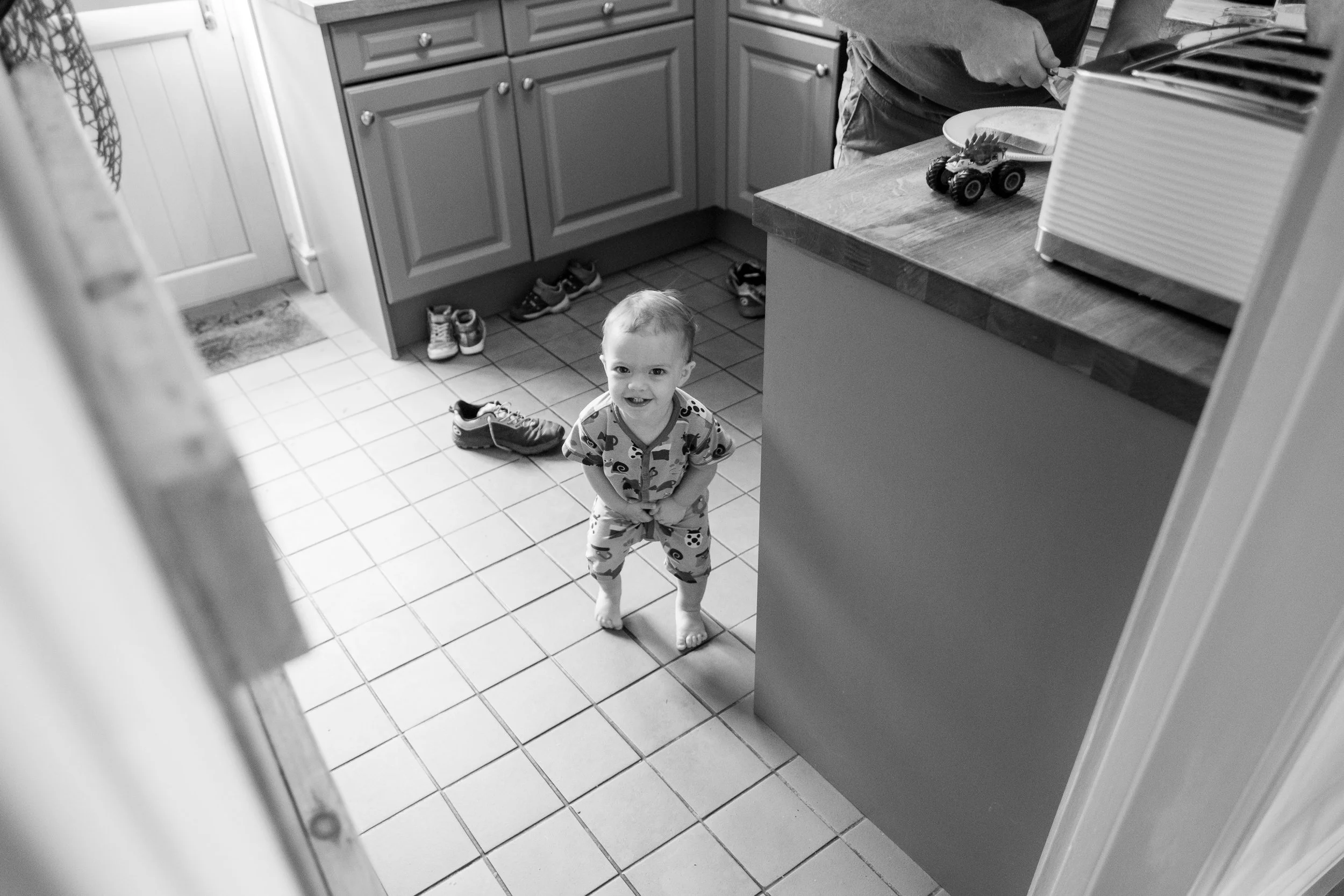 A young child standing in a kitchen, looking up and smiling, wearing pajamas with cartoon characters. Shoes are scattered on the tiled floor and a person is seen preparing food at the counter.