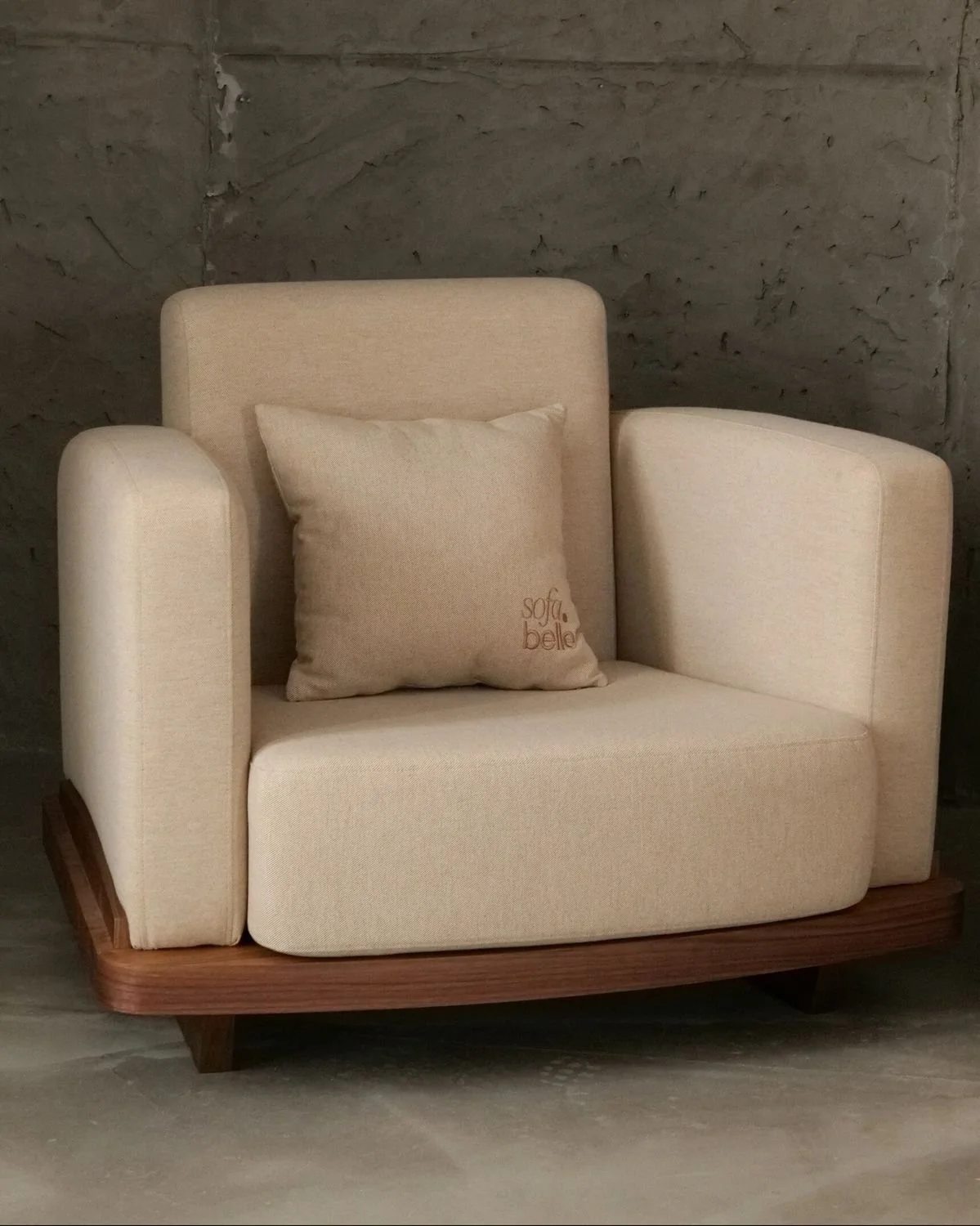 A beige upholstered armchair with wide armrests and a matching pillow that has the words 'sofa belle' embroidered on it, placed on a wooden base against a concrete block wall.