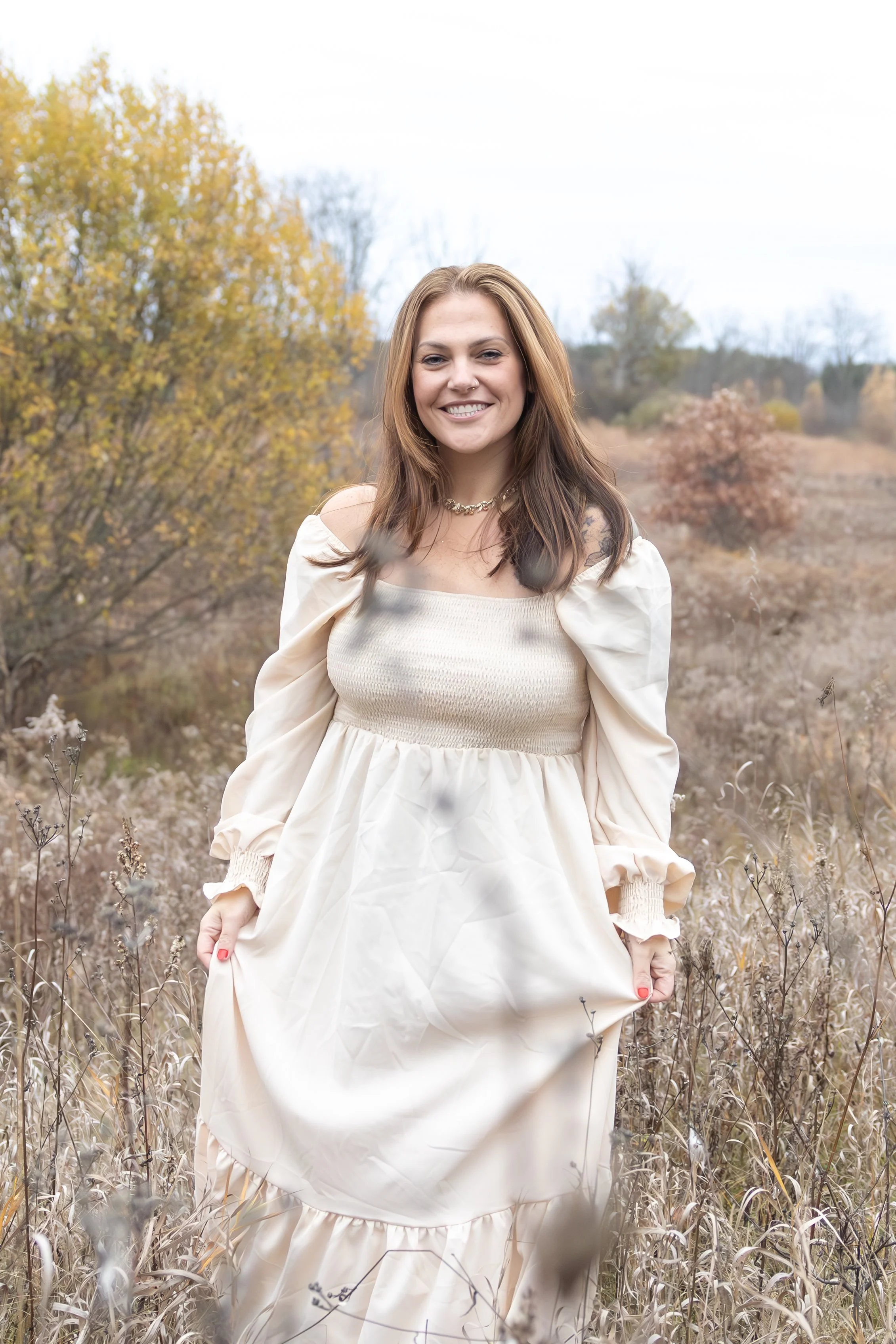 A woman with brown hair wearing a cream-colored dress with puffed sleeves standing in a field of dry grass and shrubs with trees showing fall colors in the background.