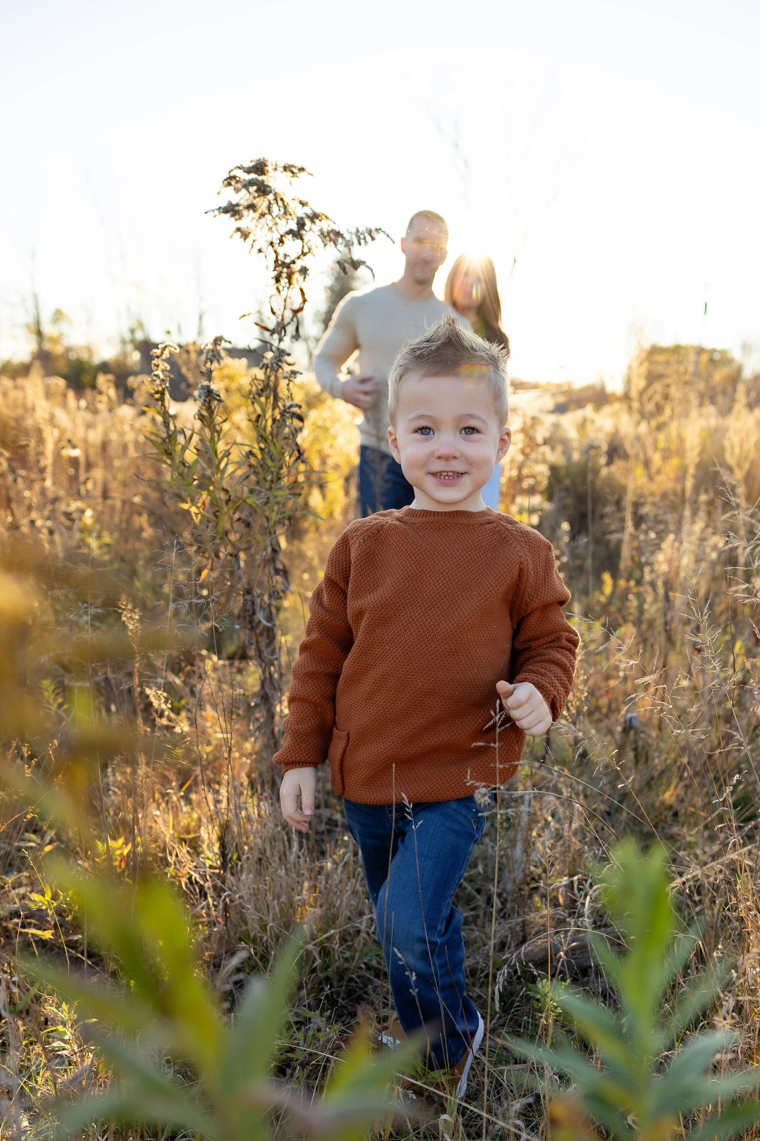 A young boy smiling and walking through tall grass in a field during golden hour, with a man and woman in the background.