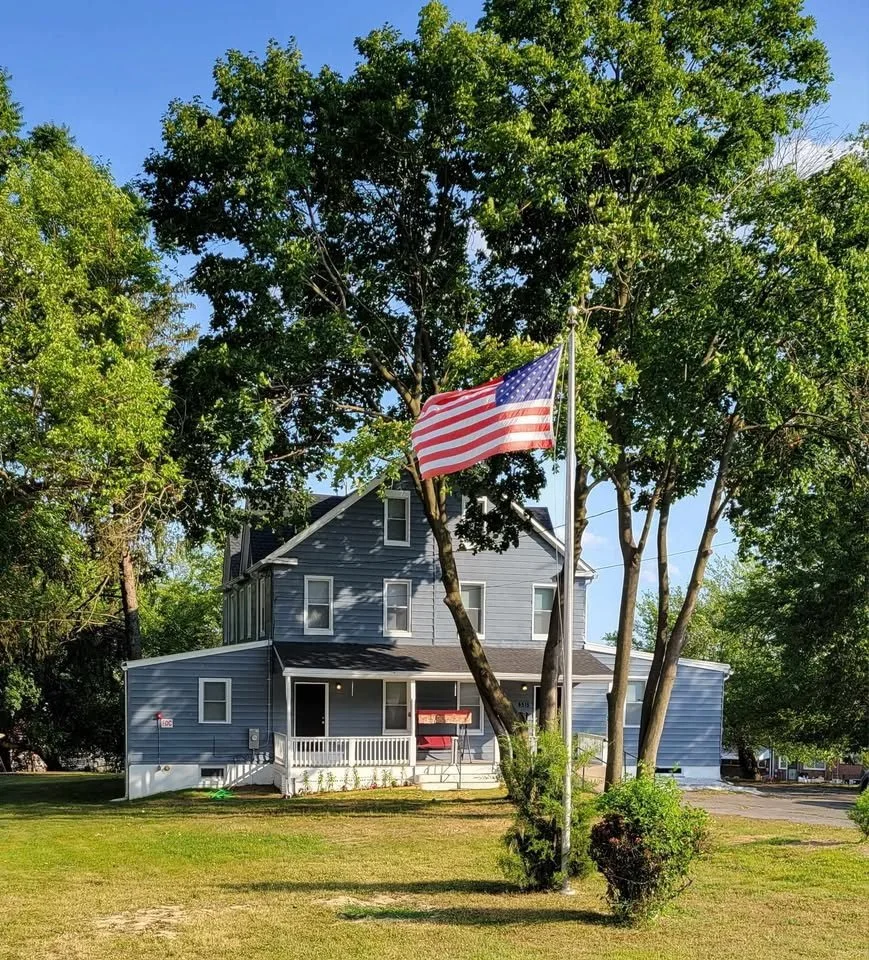 A two-story blue house with white trim and a porch, surrounded by green trees and grass, with an American flag on a flagpole in front.