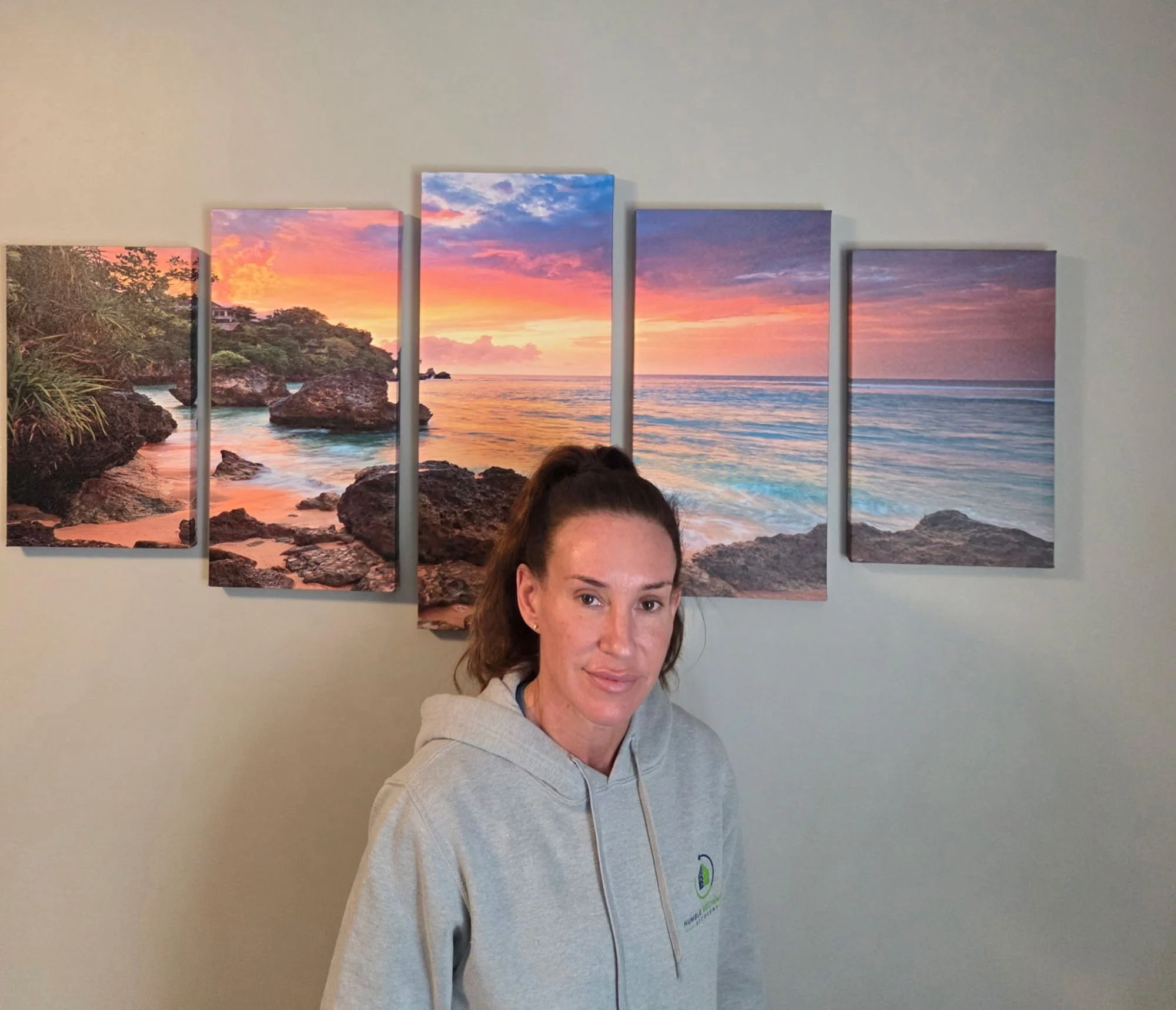 A woman with brown hair in a ponytail wearing a gray hoodie standing in front of a five-panel coastal sunset picture on a beige wall.