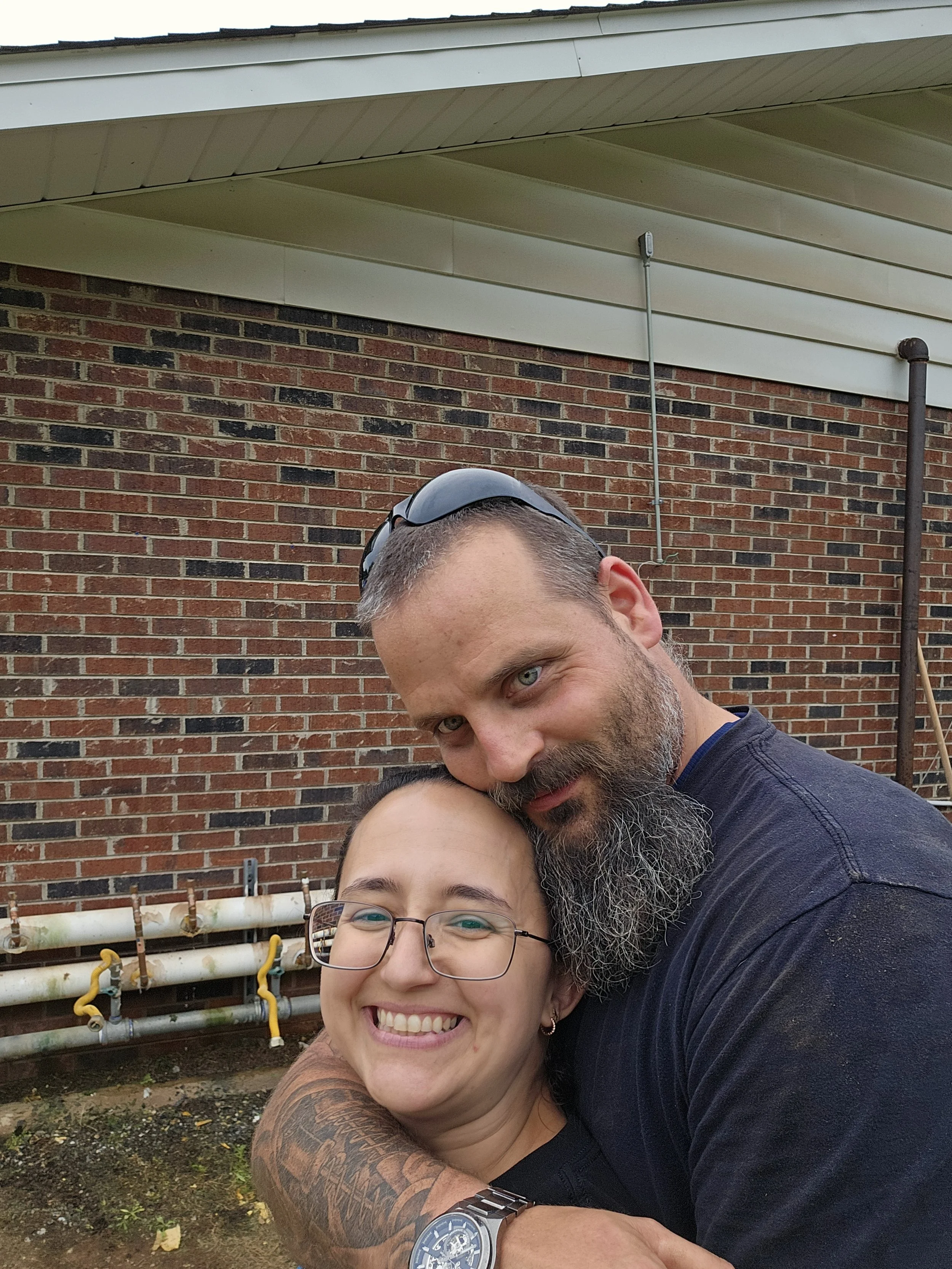 A man with a beard and tattoos hugging a woman with glasses smiling outdoors in front of a brick building.