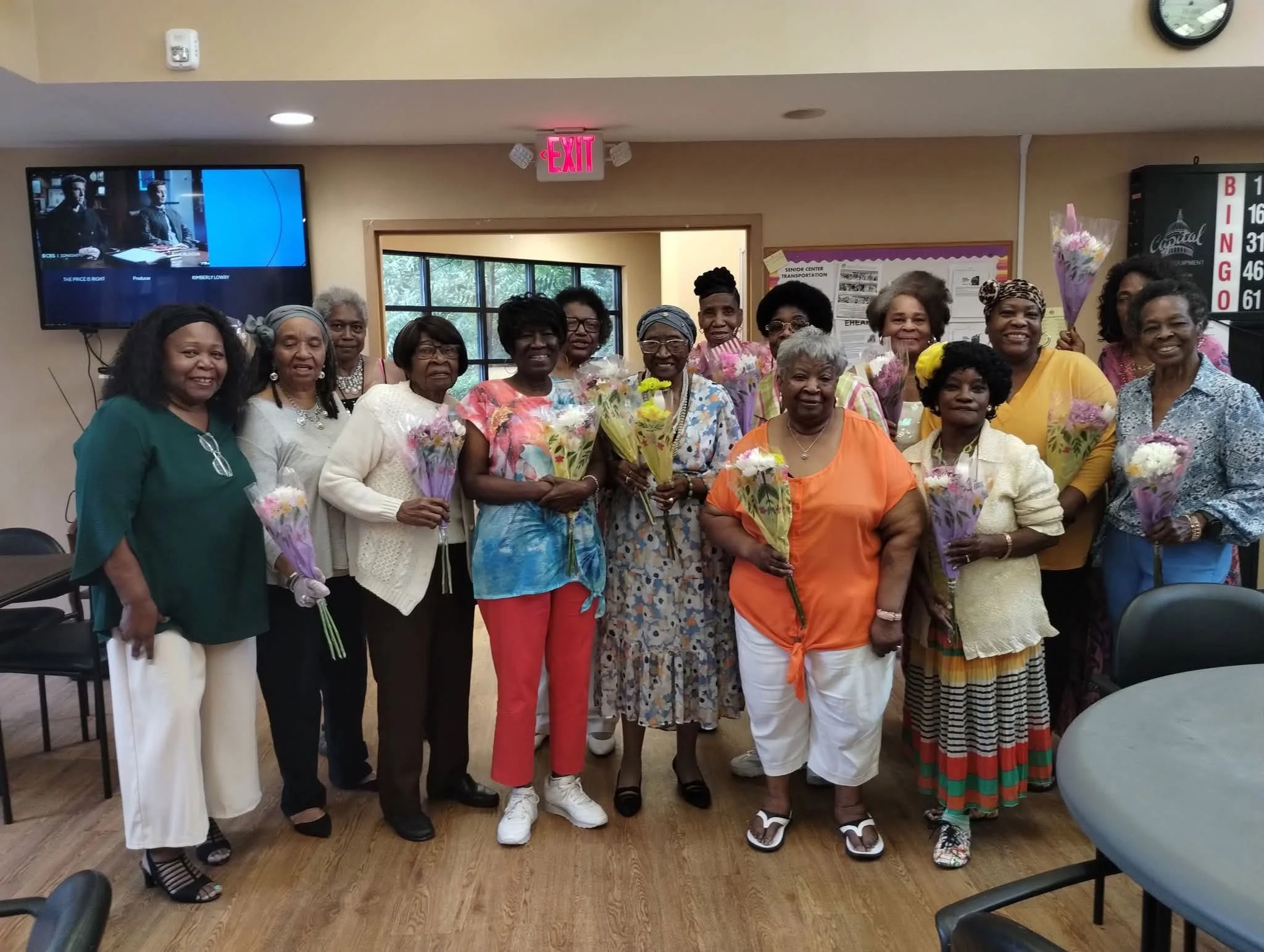 A group of women gathered indoors, holding bouquets of flowers, smiling for the photo.