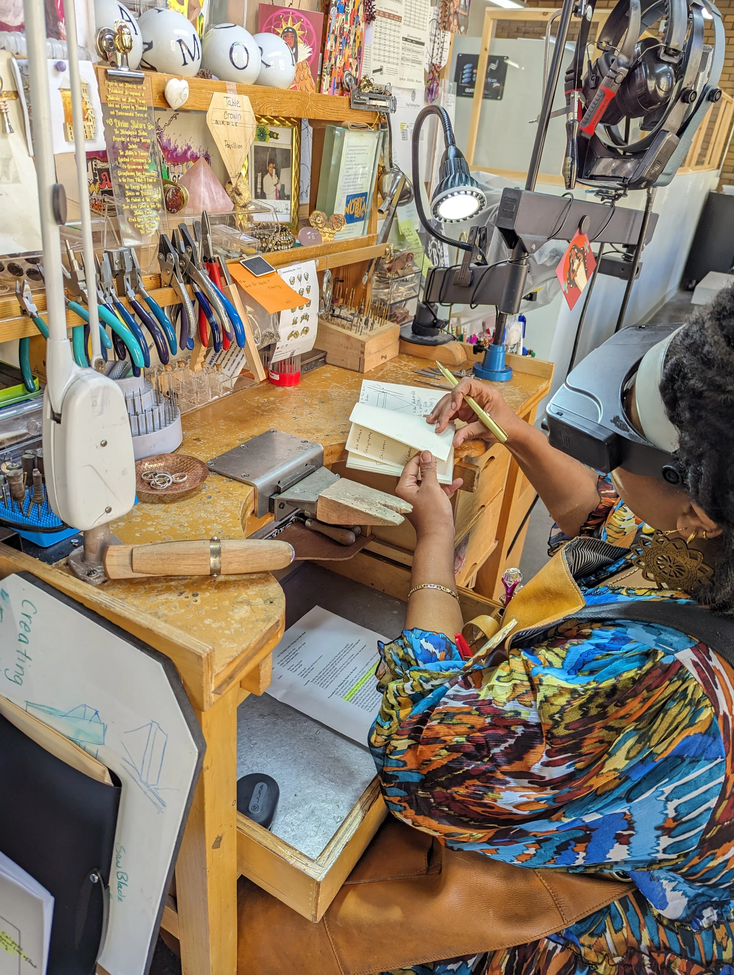 A person wearing a microscope and writing in a notebook at a jewelry workshop surrounded by various tools, jewelry, and supplies.