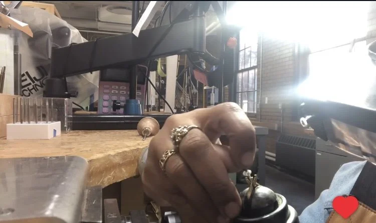 Close-up of a person's hand with rings, pressing a button on a vintage rotary phone on a cluttered wooden desk in a bright room with sunlight coming through windows.