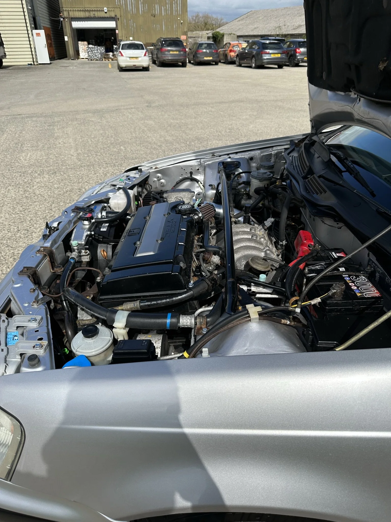 Open hood of a silver car revealing engine components in a parking lot with several other cars and industrial buildings in the background. HONDA CIVIC MB6