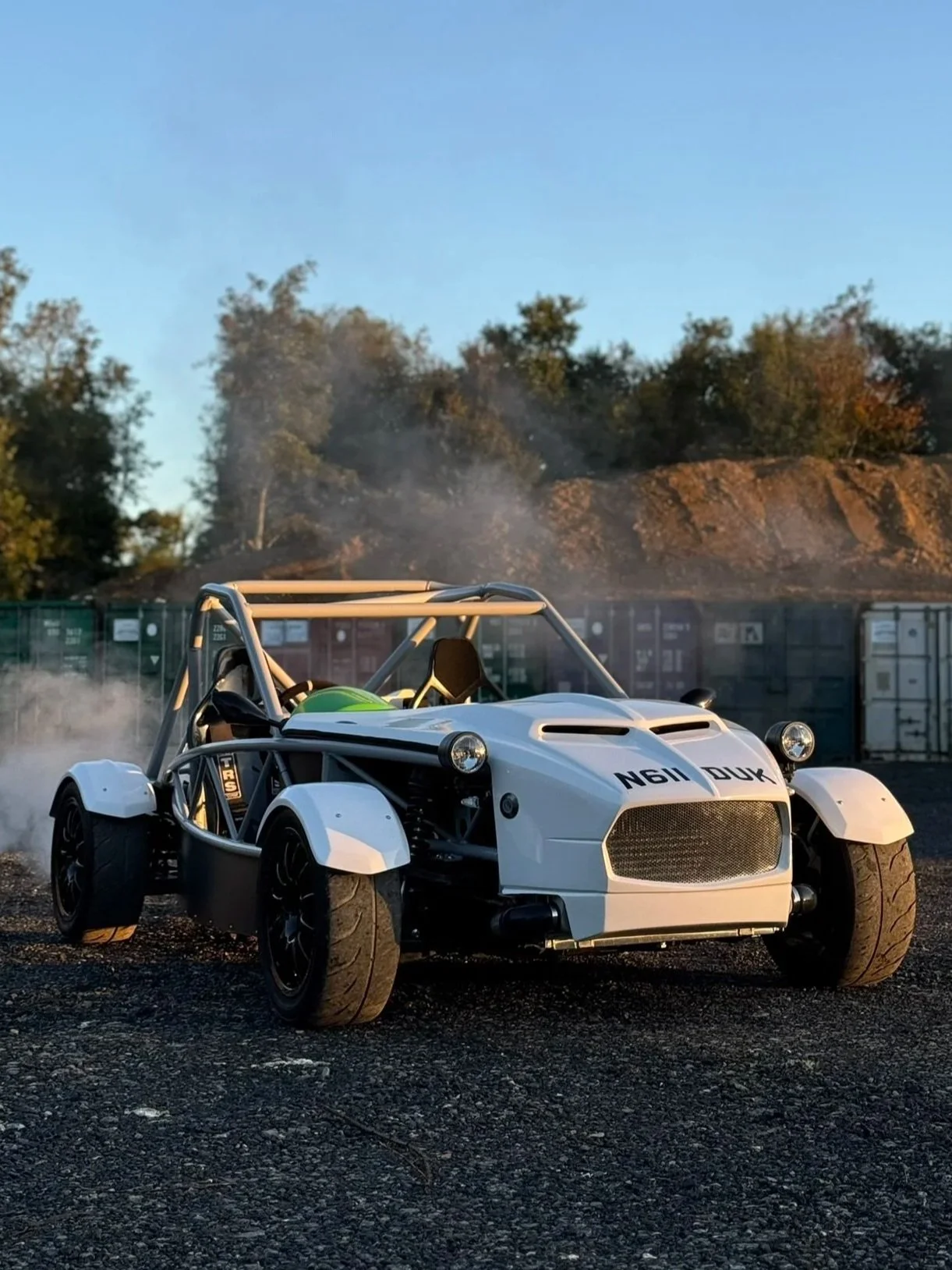 A white open-wheel race car on gravel ground with smoke emitting from the tires, set against a background of trees and a blue sky. MEV exocet