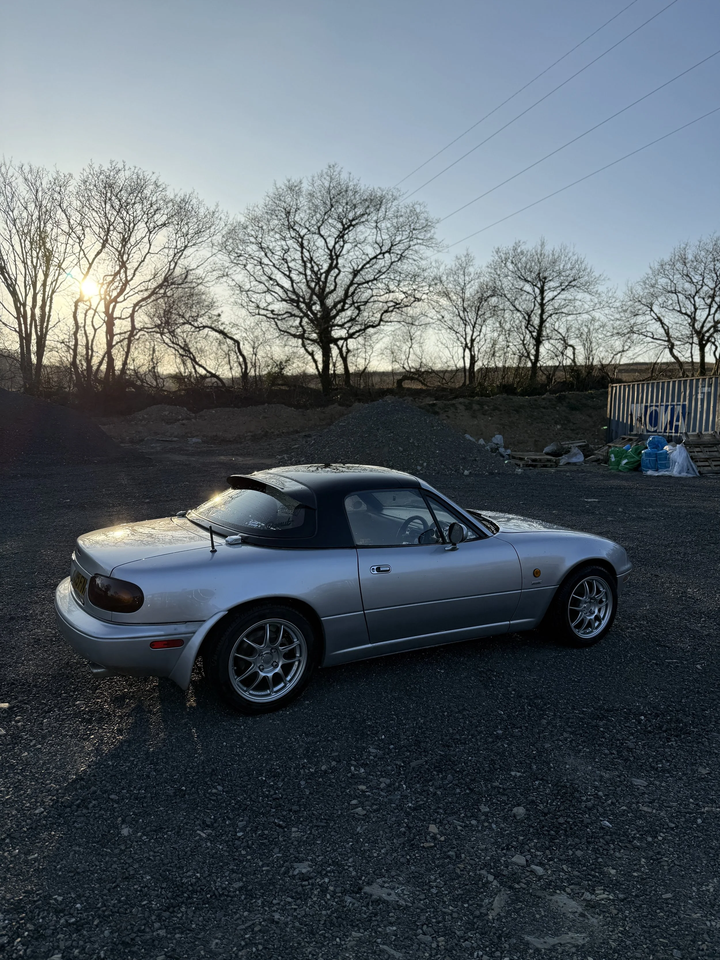 A silver convertible sports car parked on gravel with leafless trees and a sunset in the background. Mazda Miata Mx5 