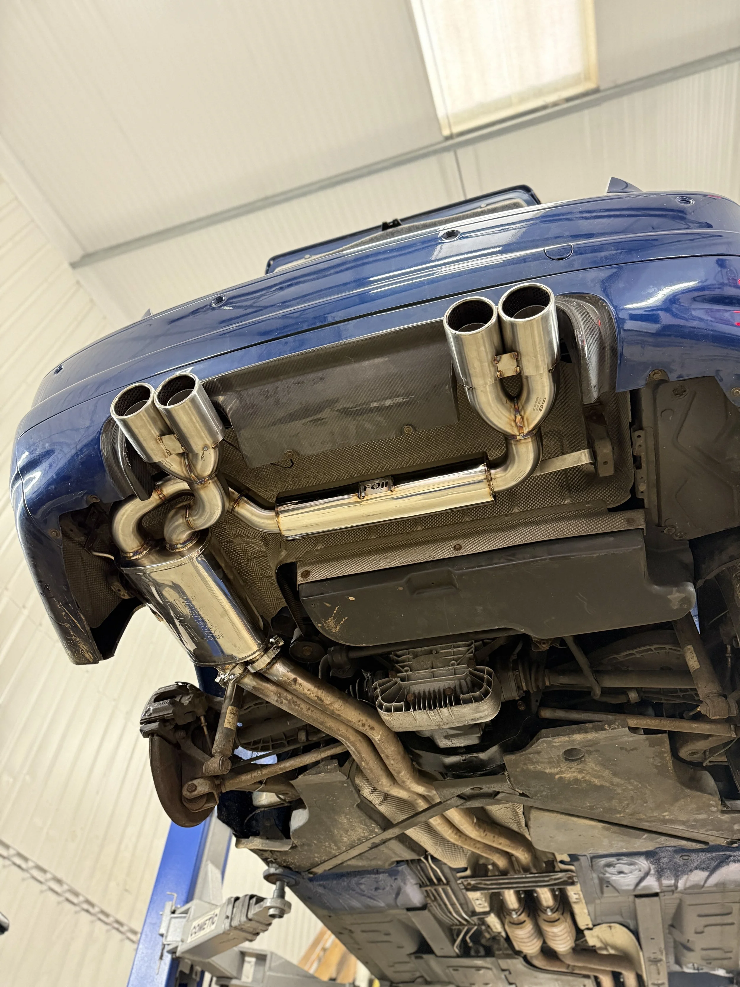 Underneath view of a blue sports car on a lift showing custom exhaust system with dual exhaust pipes. E46 BMW M3
