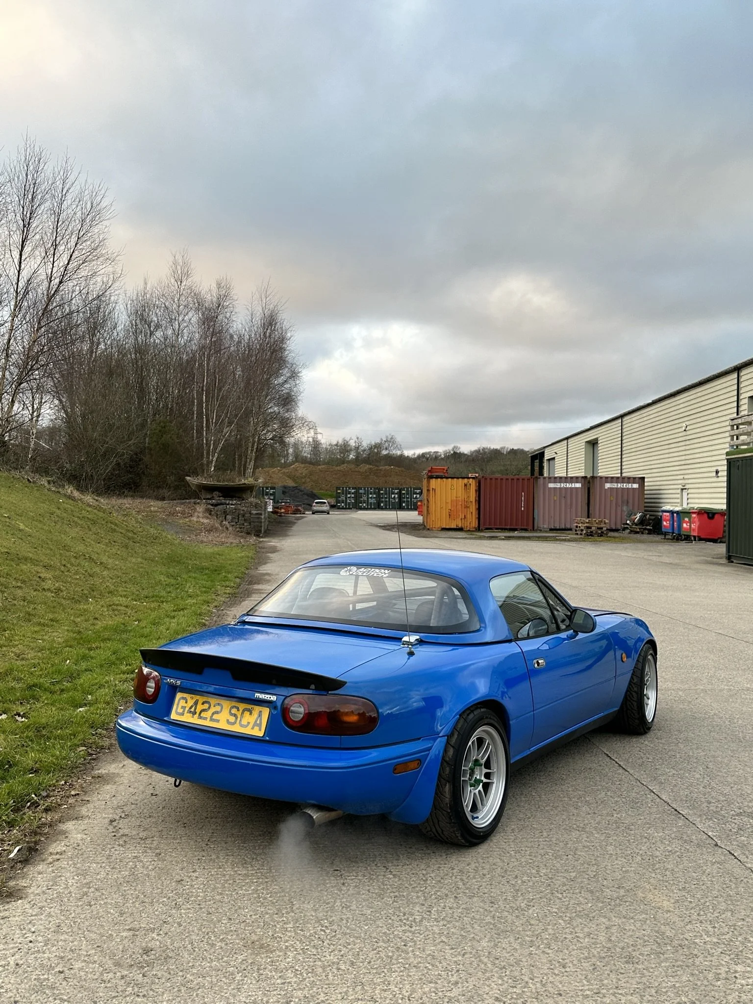 Blue Mazda MX-5 Miata sports car parked on pavement near grassy area with trees and industrial building in background.