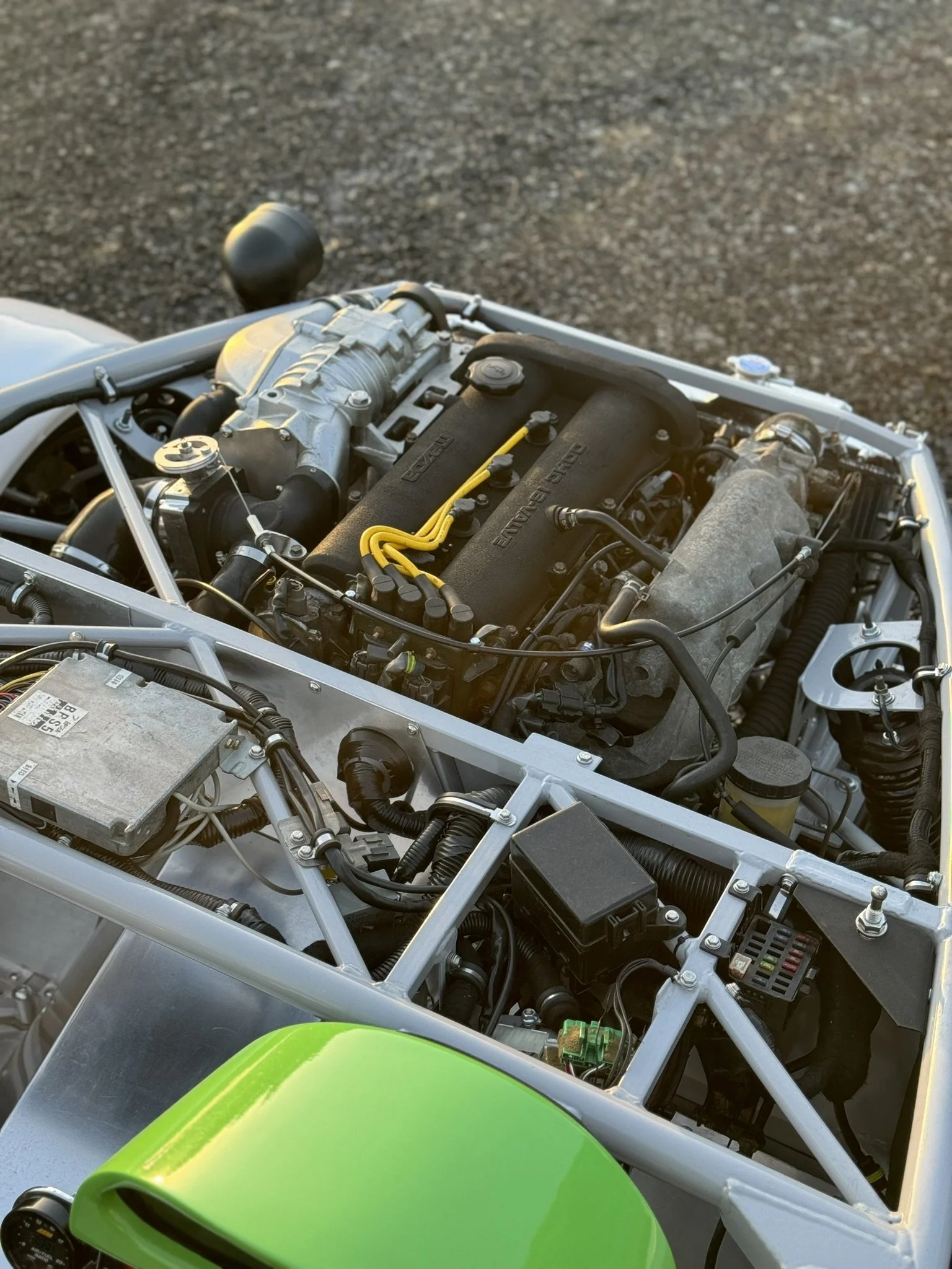 MEV Exocet Close-up photo of a car engine with yellow spark plug wires and a green wheel in the foreground, set on a paved surface.