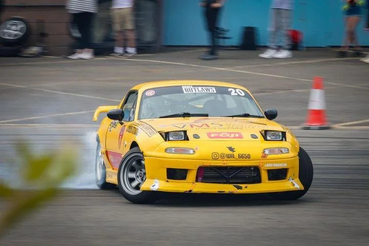 Yellow drift car making a turn, emitting smoke from the rear tires, with onlookers and traffic cones in the background. Mazda Miata mk1 eunos 